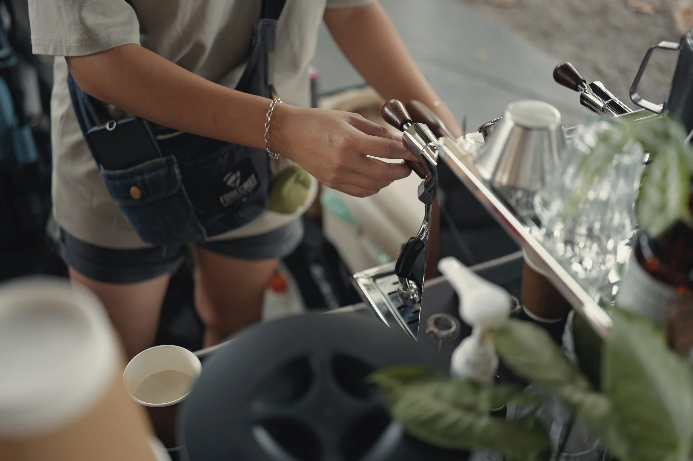 A person prepares a drink beside stacked cups and metal utensils at a café counter.