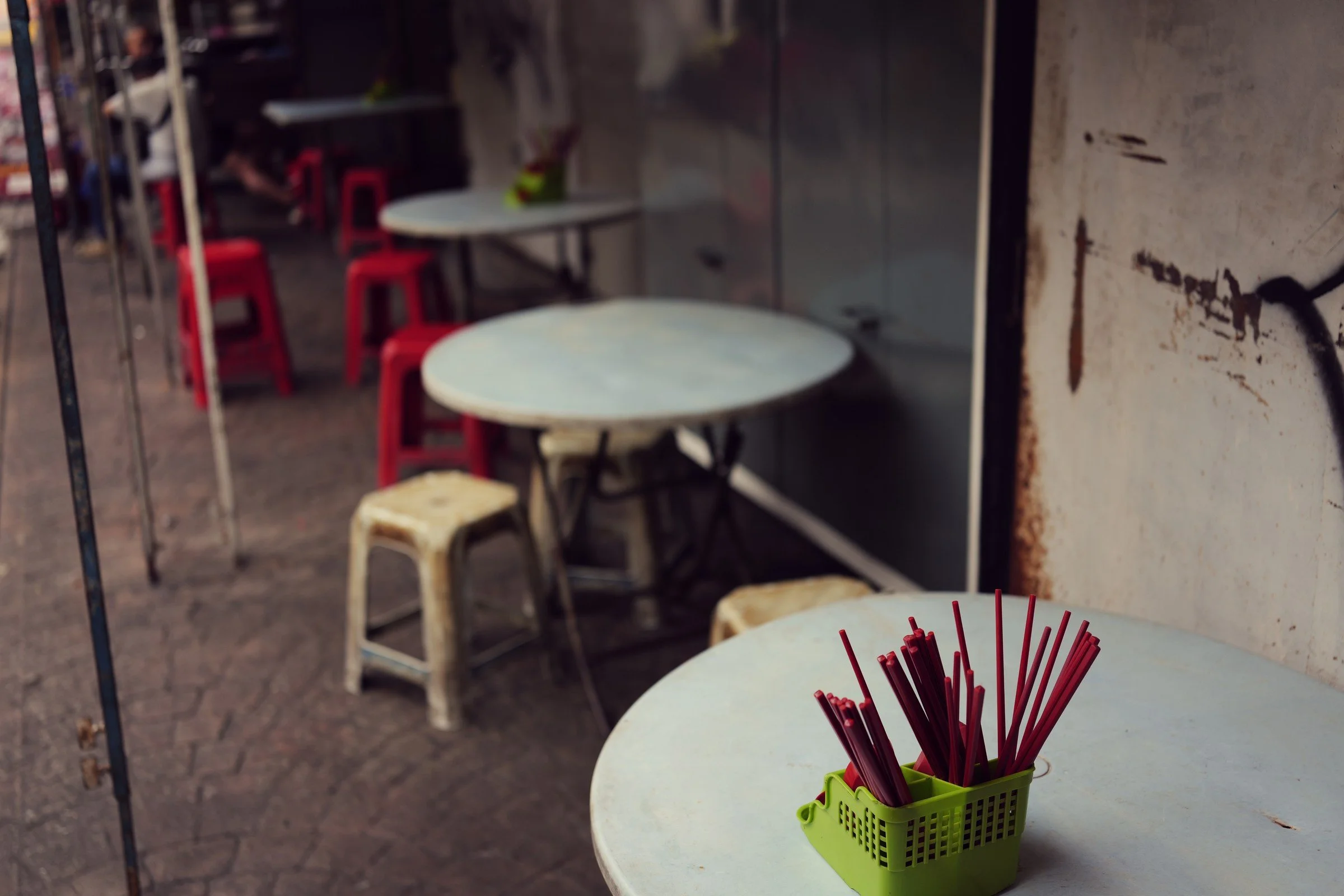 A cup of red chopsticks sits on a small table beside an old shop wall.