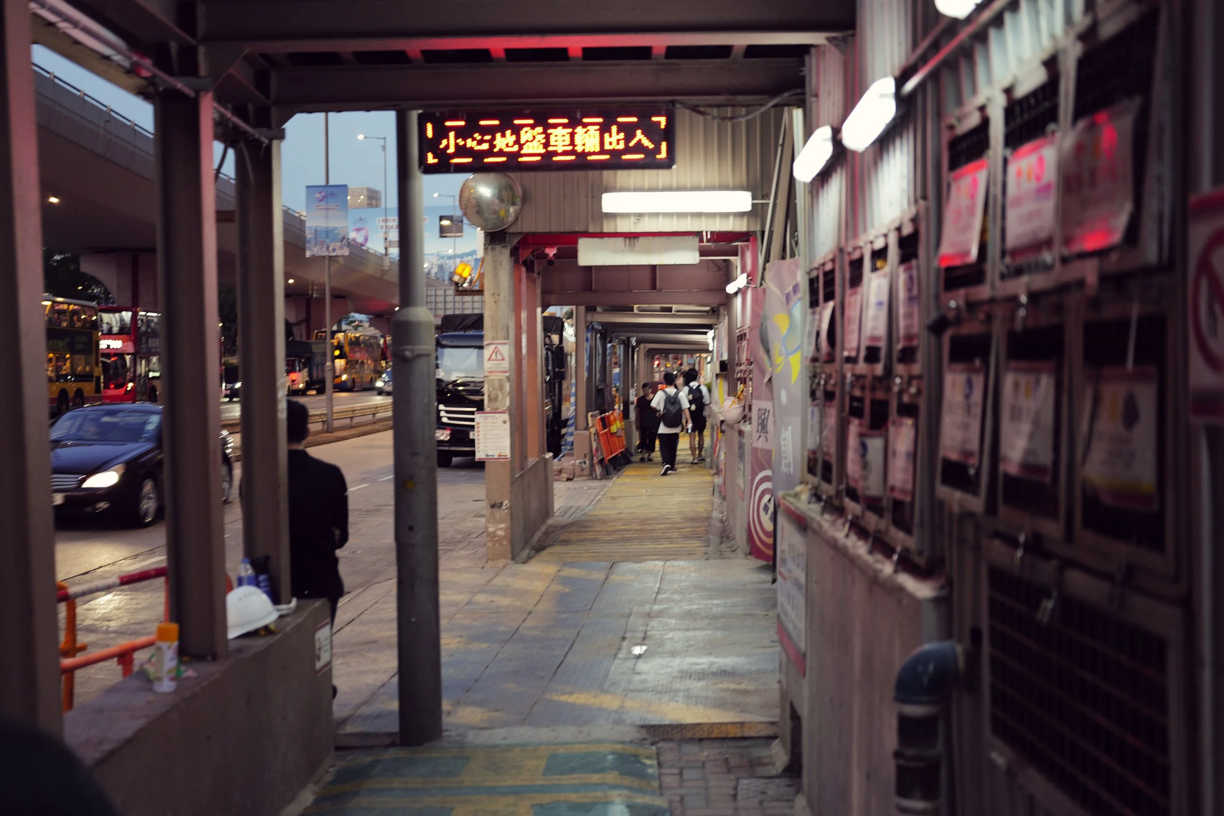 Urban sidewalk scene with people walking, parked cars on the street, and a digital sign with Chinese characters overhead. Sidewalk has newspaper dispensers and construction helmets.