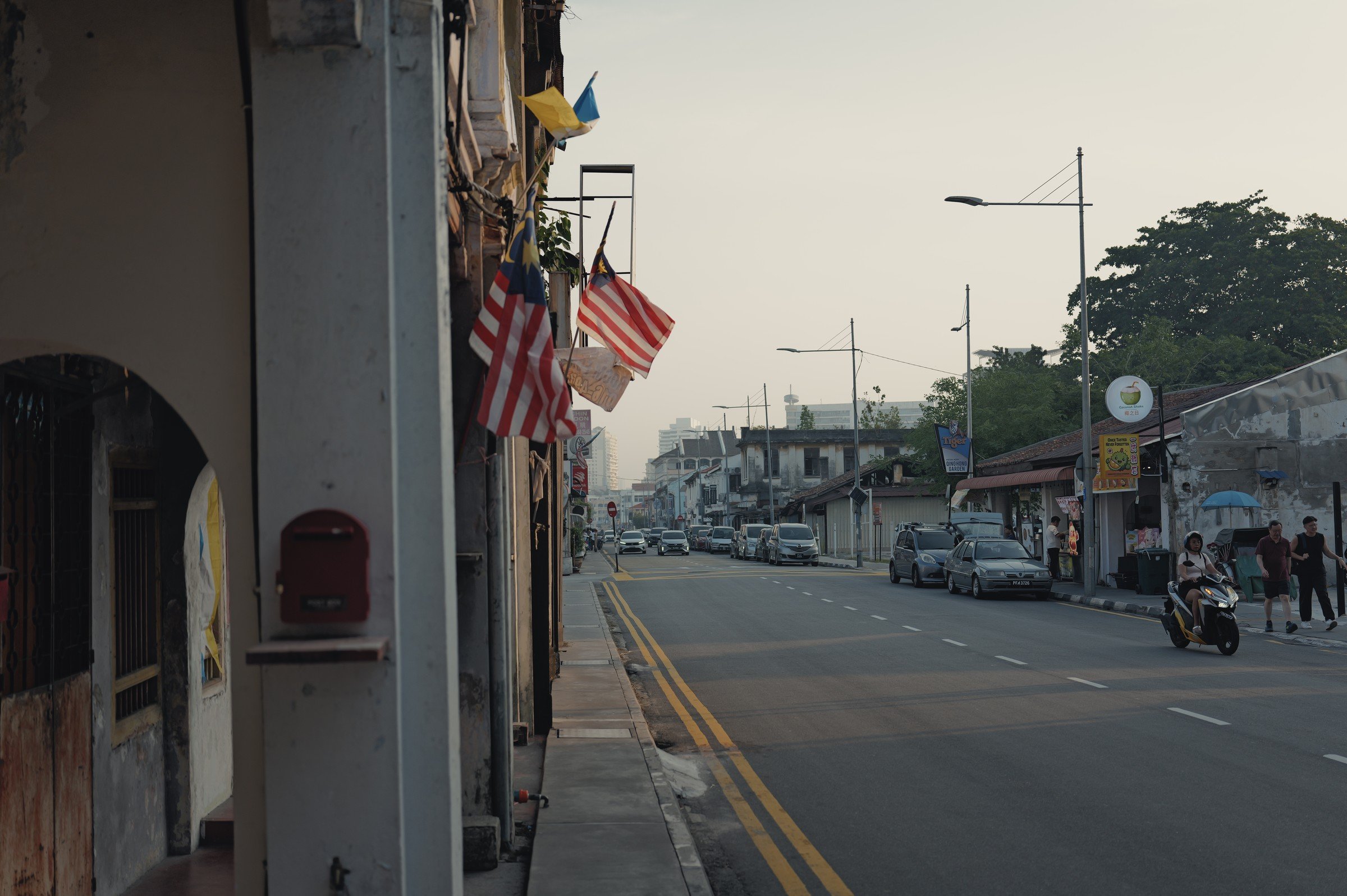 A quiet street lined with low shophouses and Malaysian flags in soft afternoon light.