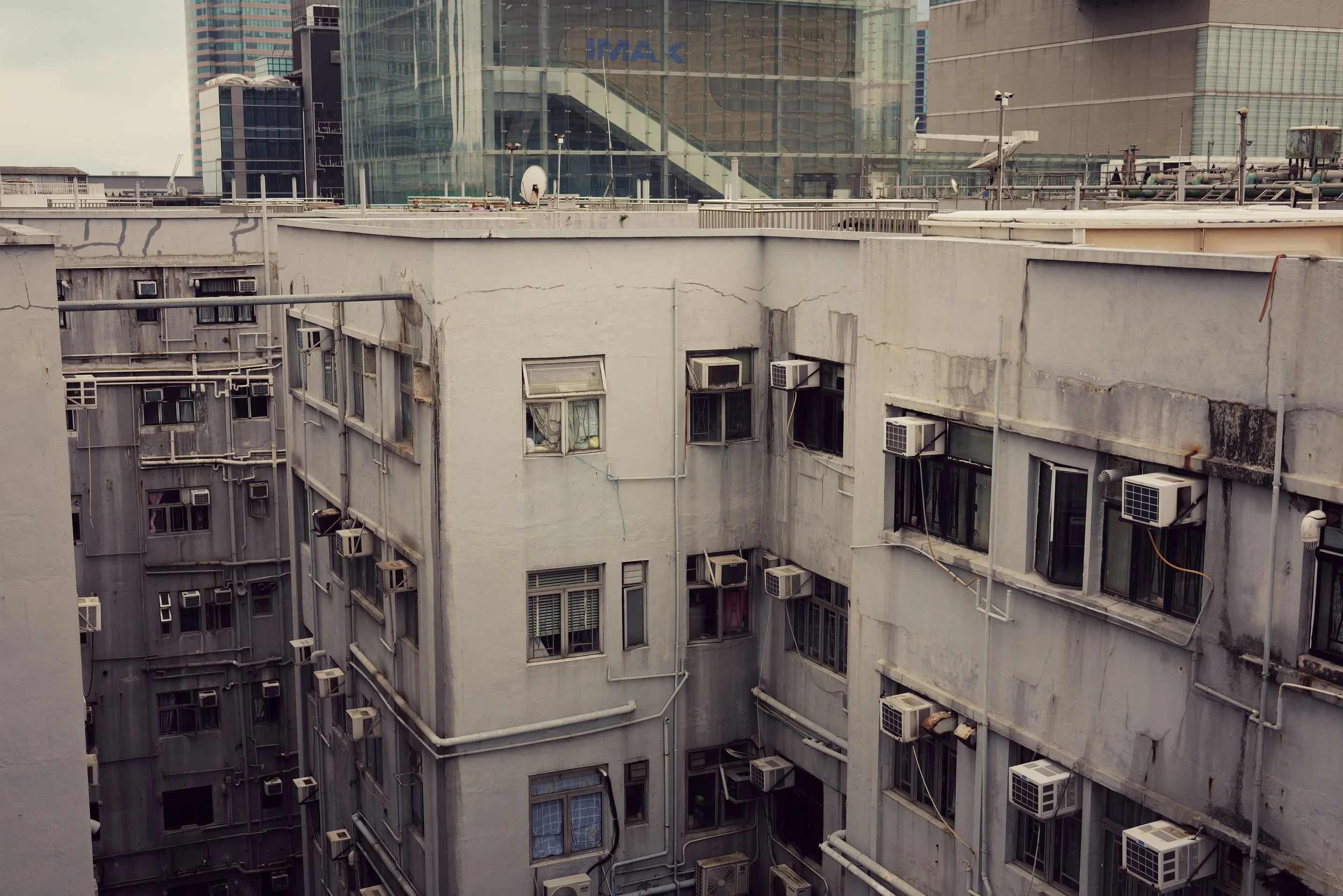 View of city buildings with rooftop air conditioning units, pipes, and weathered paint on the structures in the foreground, and modern skyscrapers and a glass mall in the background.