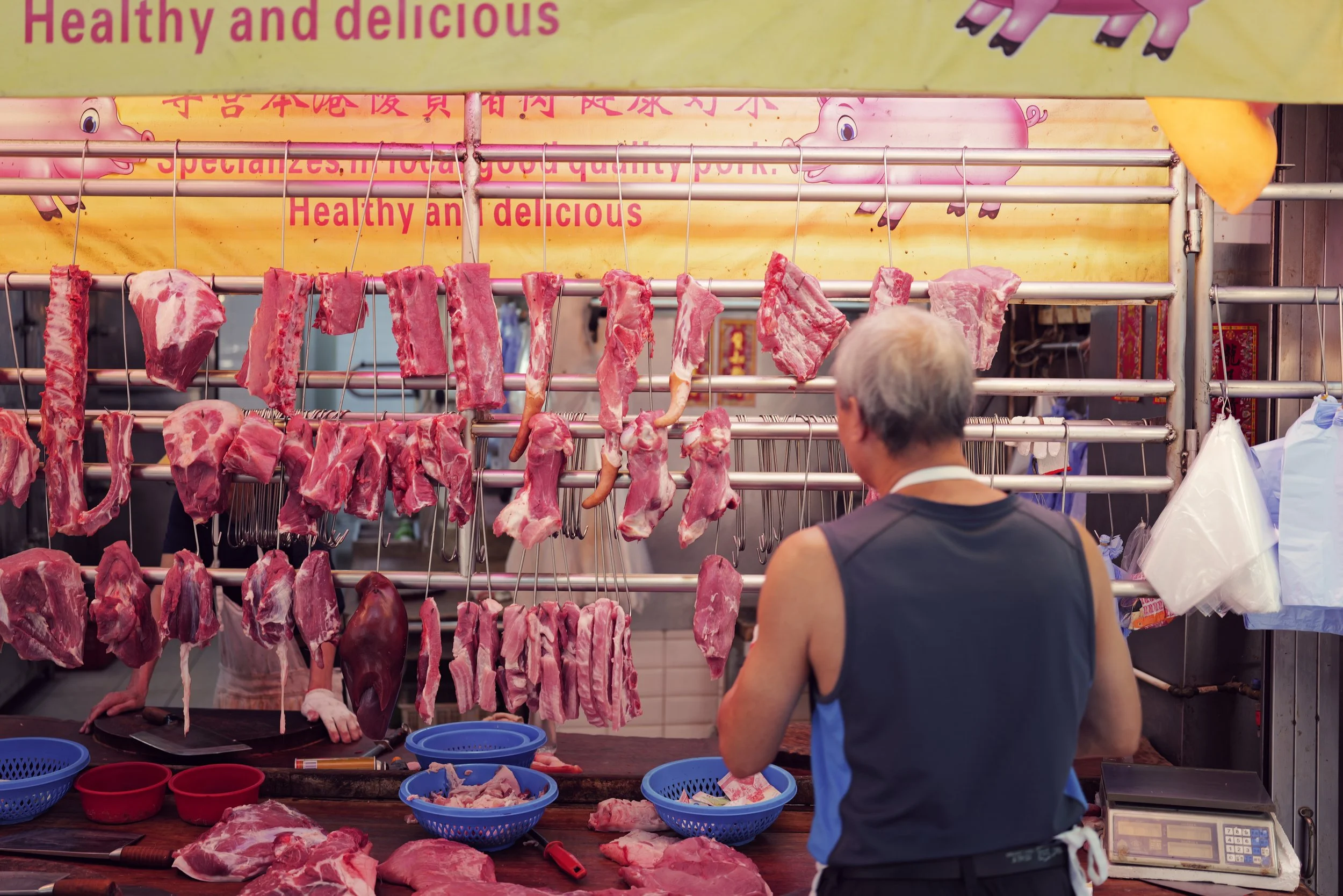 A man selling fresh cuts of meat at a market stall, with meat hanging on hooks and others on a table in front, and a colorful sign overhead.