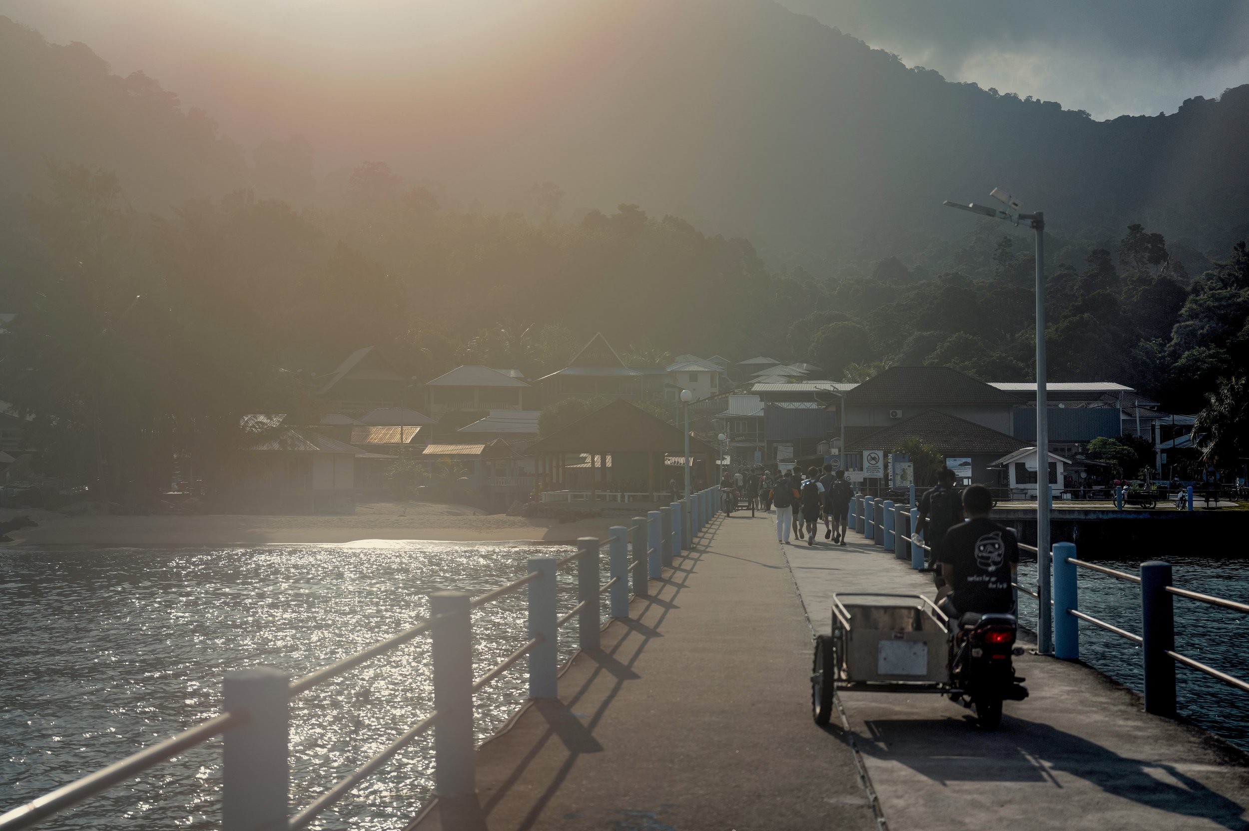 People walking on a waterfront pier with houses and green hills in the background during sunset.