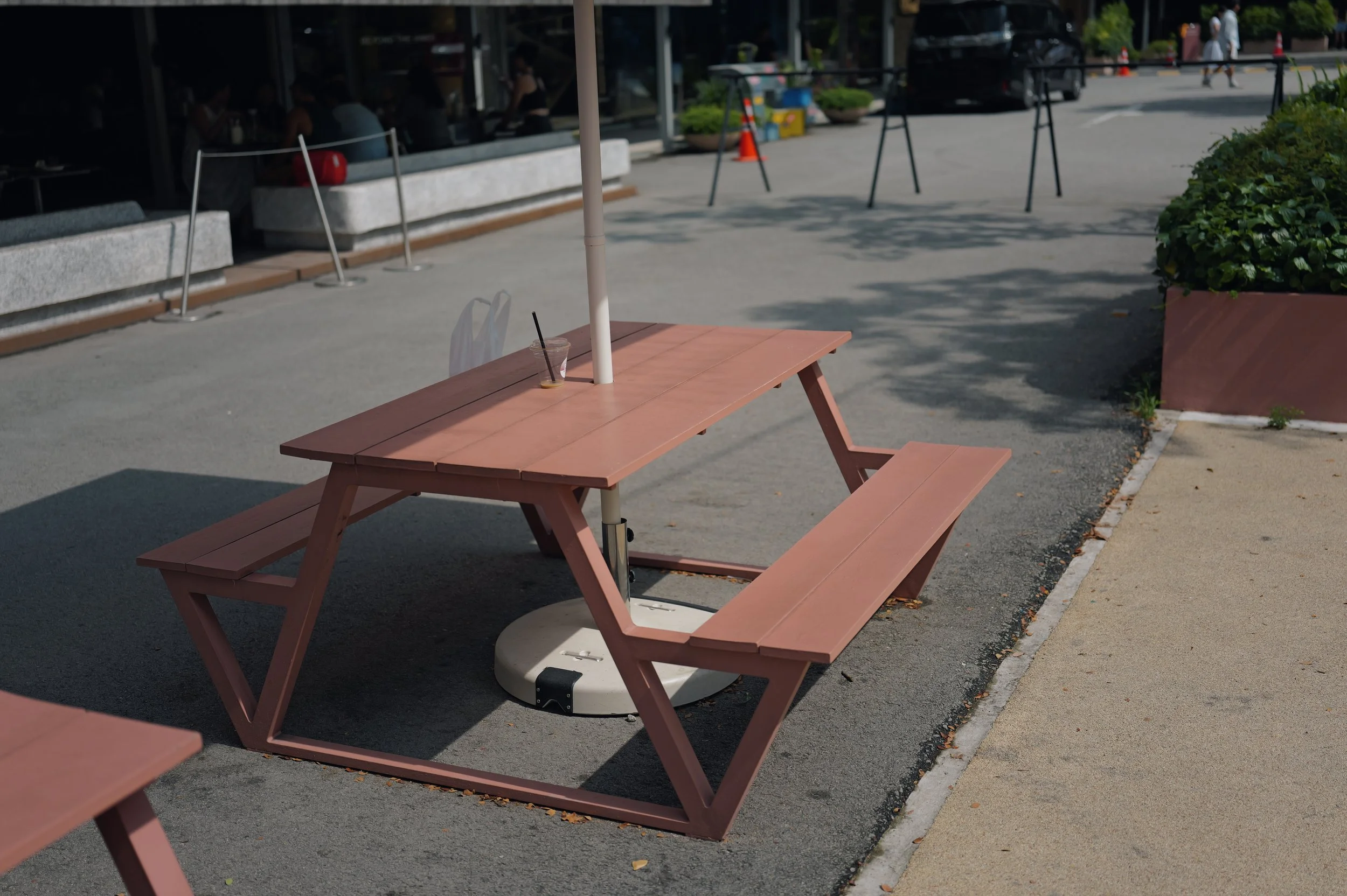 Empty pink picnic table with a white umbrella pole in a shopping plaza outdoor seating area. Some customers visible inside the building in the background, with a few outdoor traffic cones and parking spaces outside.