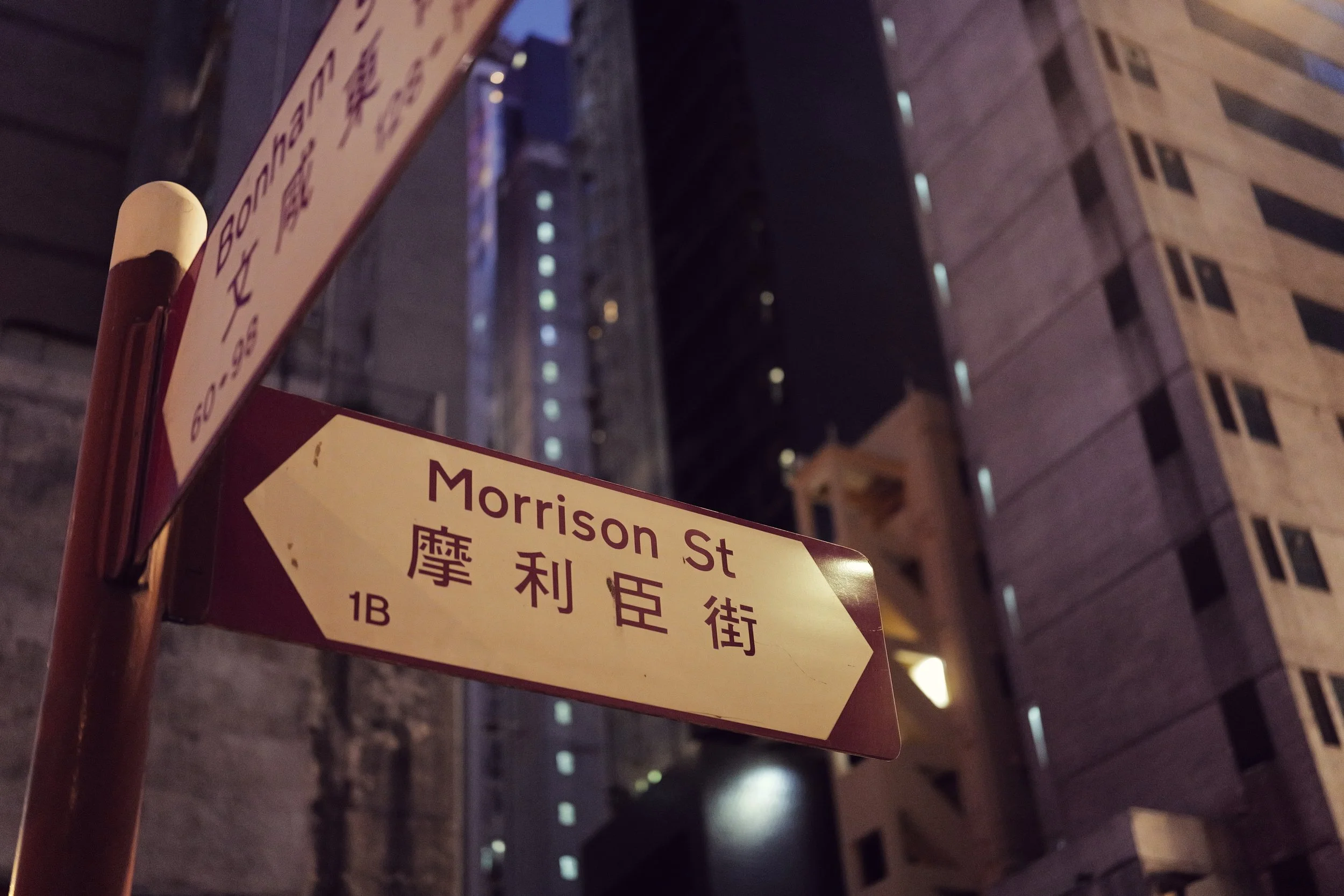 Street sign at night with the text 'Morrison St' in English and Chinese characters, against a background of a tall building with lit windows.