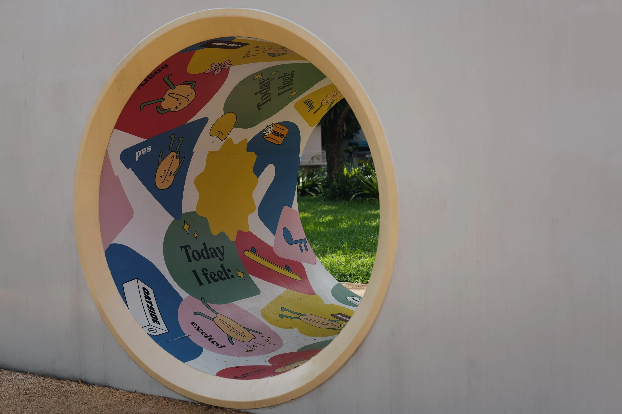 View through a circular opening in a white wall, showing a colorful poster with illustrations and text, outdoors with grass and trees in the background.