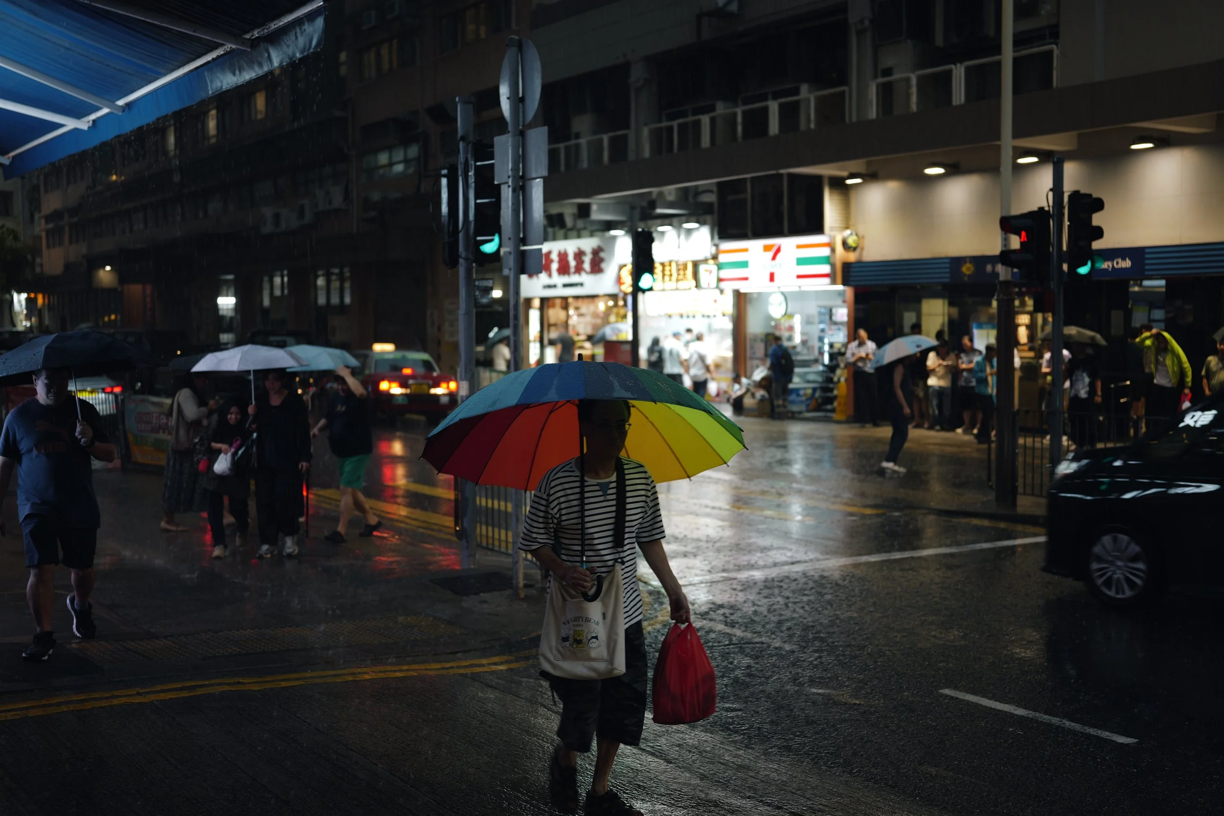 People walking on a rainy city street at night, some holding umbrellas, with storefronts and traffic lights in the background.