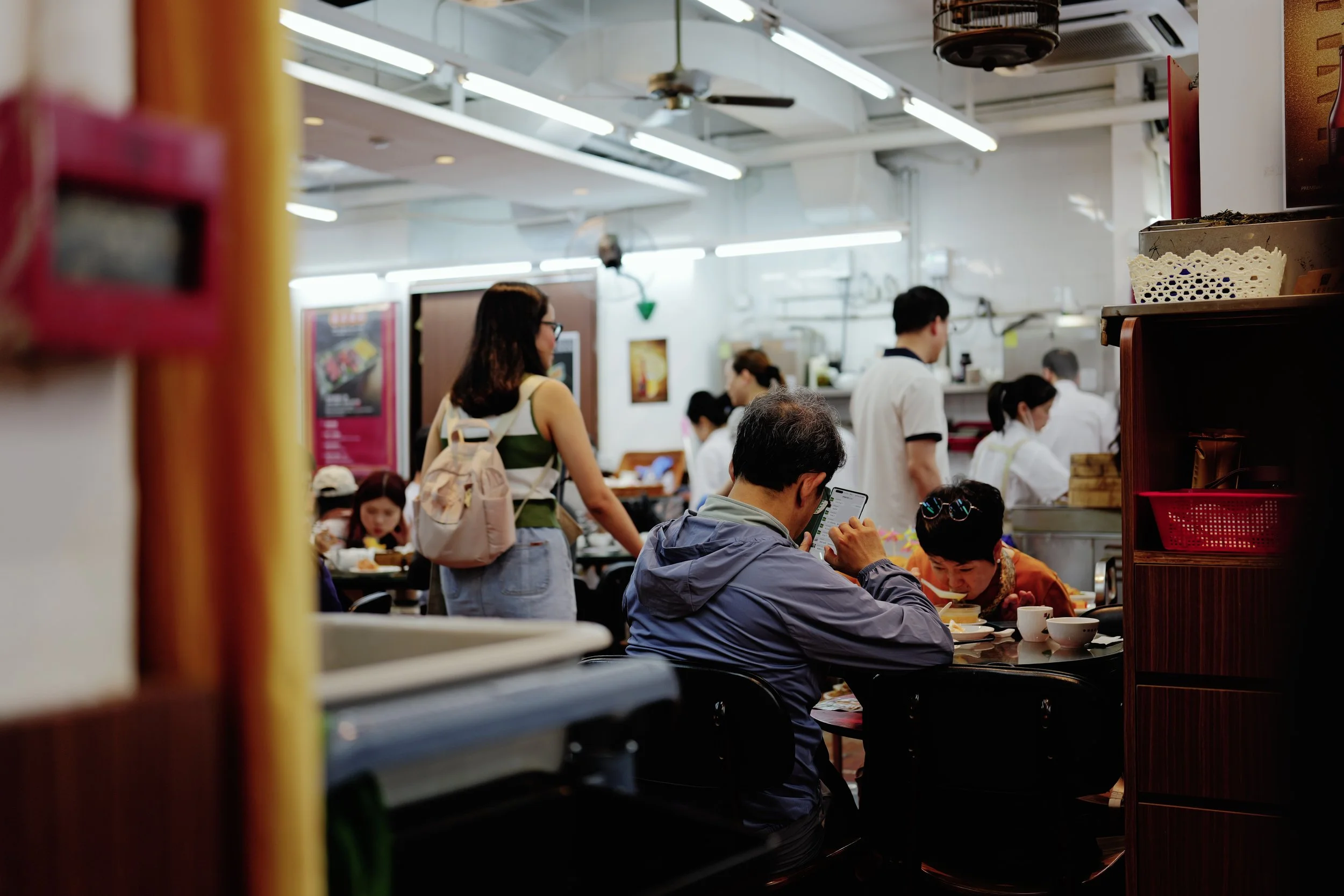 People dining in a busy restaurant, some are seated at tables eating, others are standing and ordering food, with the kitchen area visible in the background.
