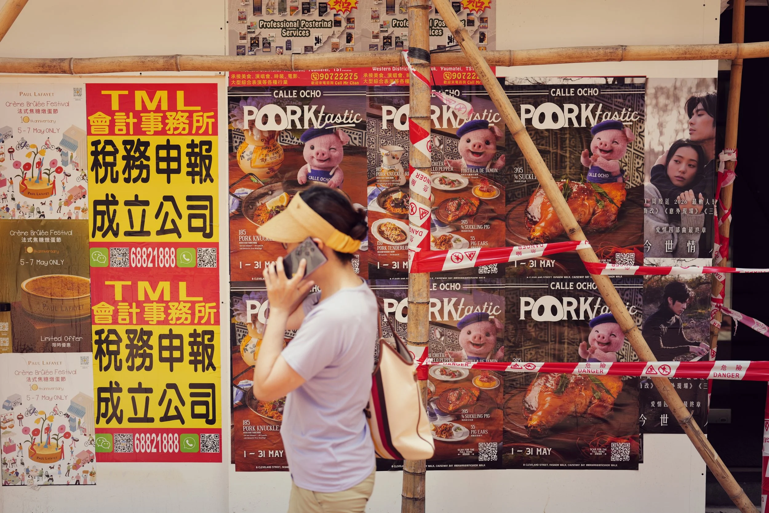Person in a white shirt and beige shorts taking a photo or selfie in front of a display of posters, which include images of pigs, food dishes, and advertising text in Chinese and English, with bamboo scaffolding in the foreground.