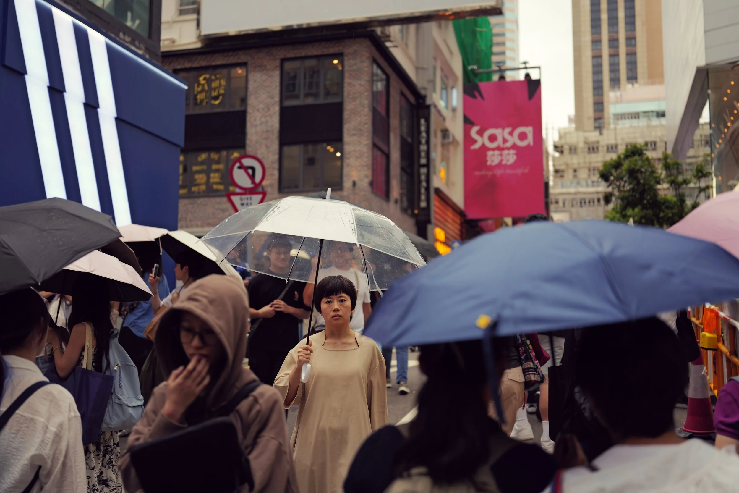 Crowd of people with umbrellas on a city street during rain, with buildings, signs, and trees in the background.