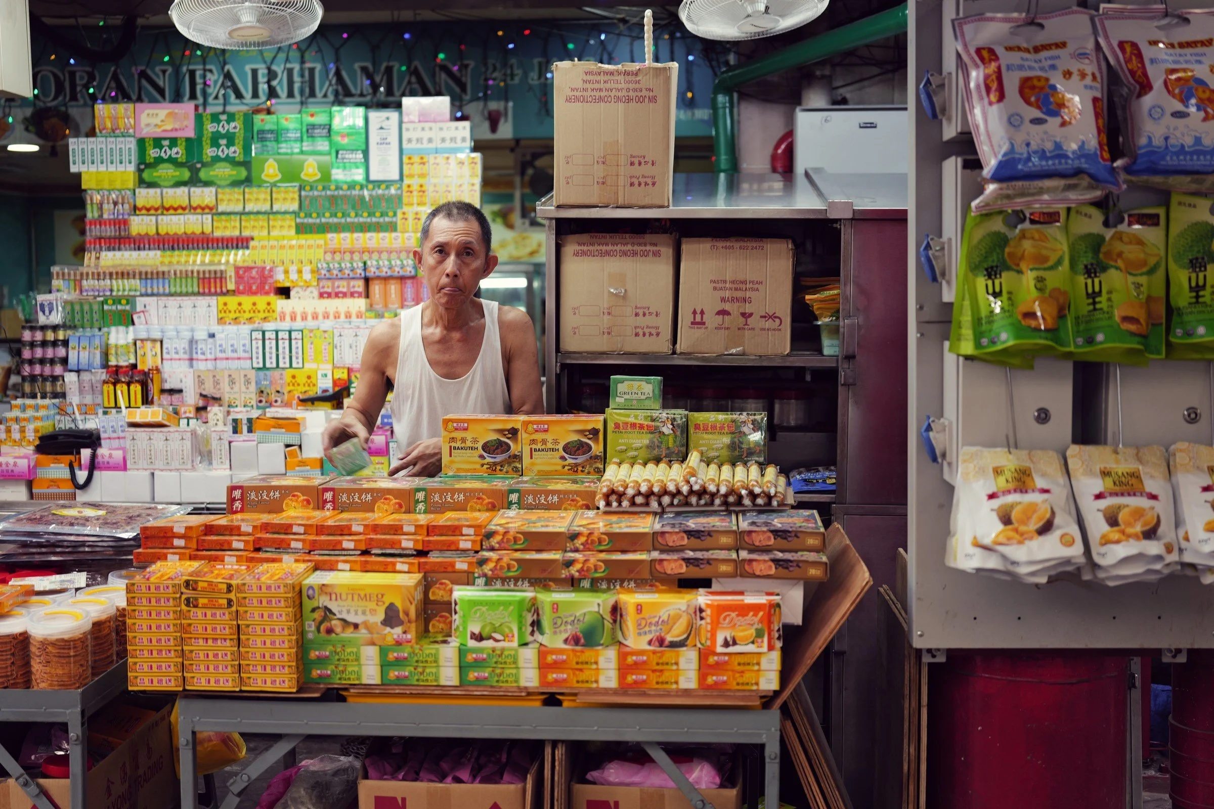 A shopkeeper sits behind a counter filled with neatly stacked small goods.