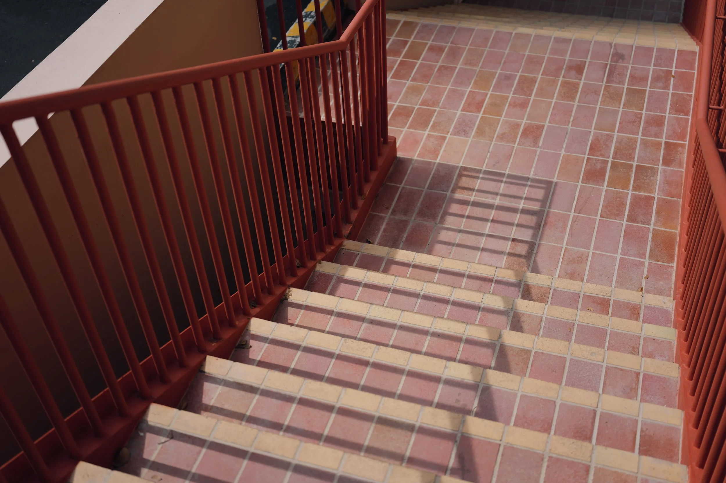 Outdoor staircase with terracotta-colored tiles and red metal railings, casting shadows on the steps.