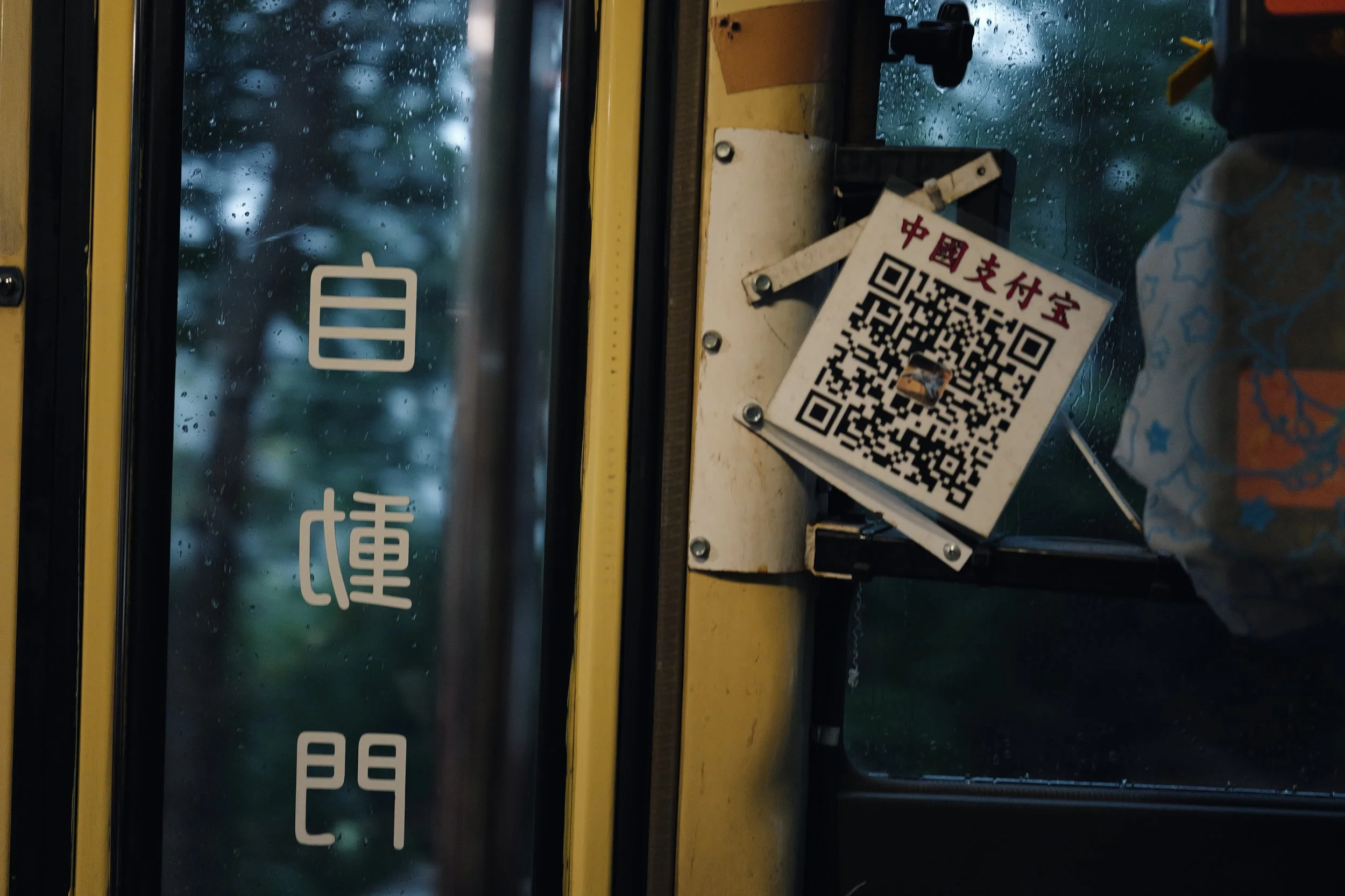 A window on a bus shows raindrops on the glass with a blue background. There is a white sticker with Chinese characters and a QR code attached to the bus window, with some metal screws and a yellow stripe visible on the bus interior.