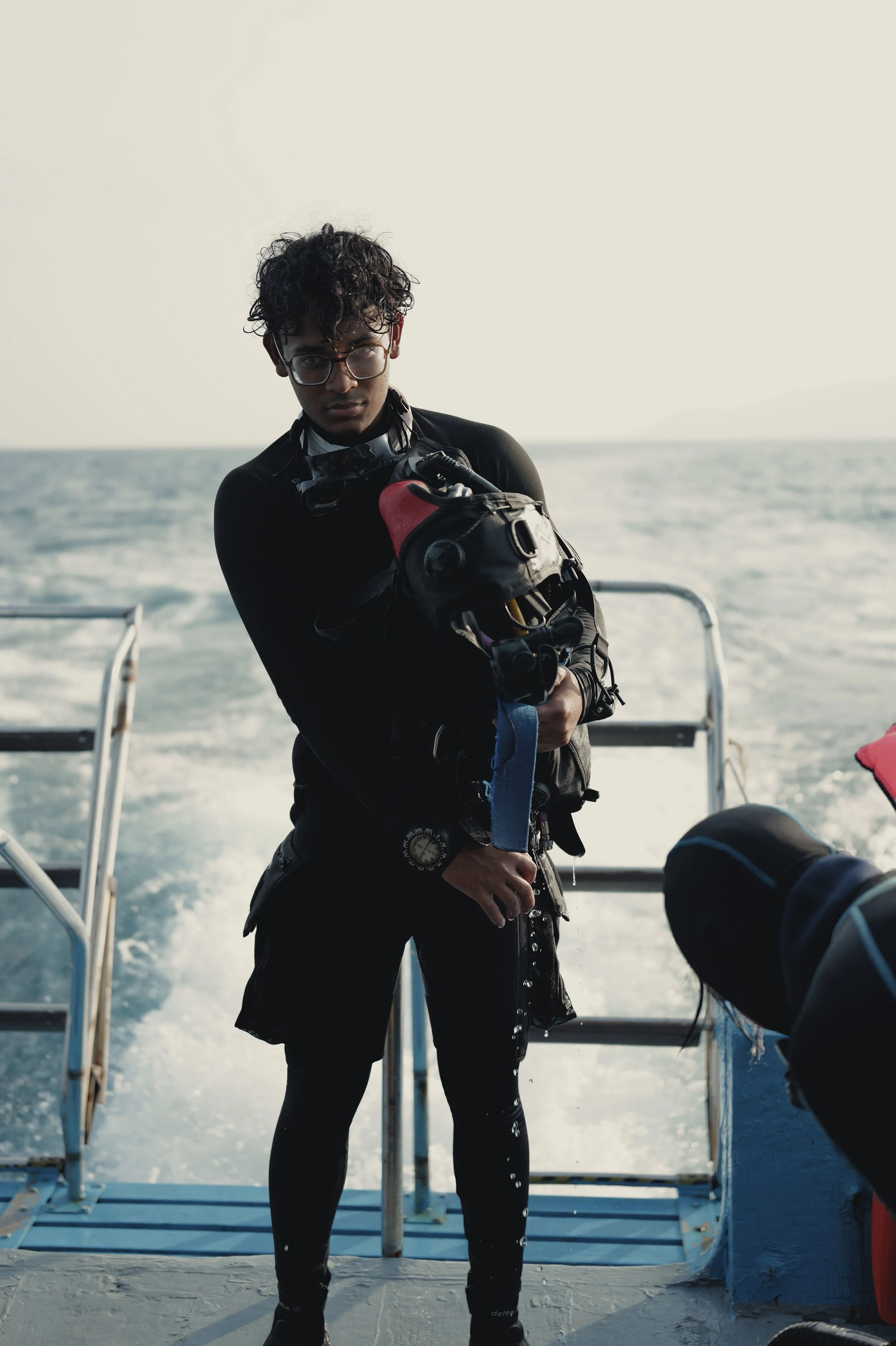 A person with curly hair and glasses wearing winter gear, holding scuba diving equipment, standing on a boat with the ocean in the background.