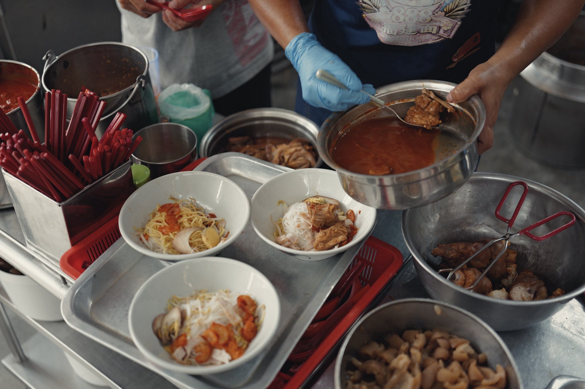 Bowls of Penang prawn noodles being assembled beside simmering broth pots at a hawker stall.