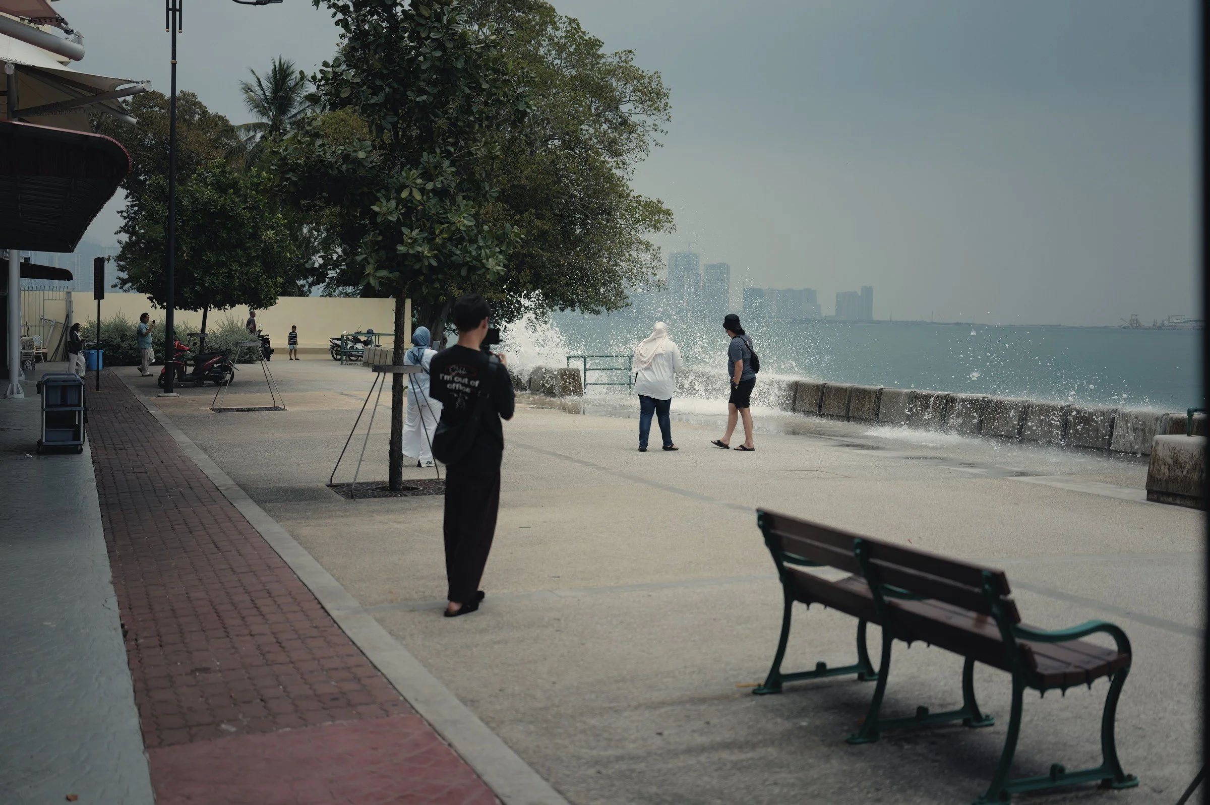 People pause on a waterfront promenade as a wave breaks over the seawall near a bench.