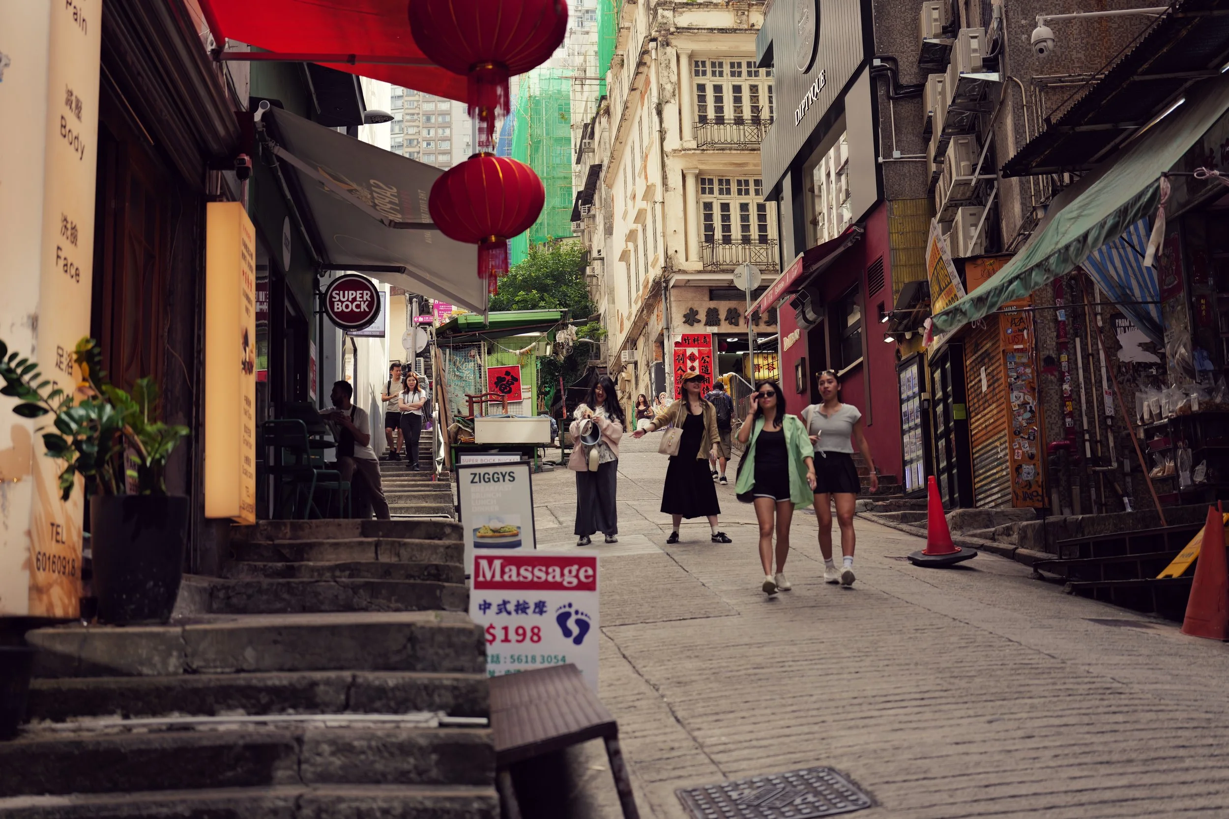 A busy city street with pedestrians walking, storefronts, red lanterns hanging, and signs advertising food and services, with high-rise buildings in the background.