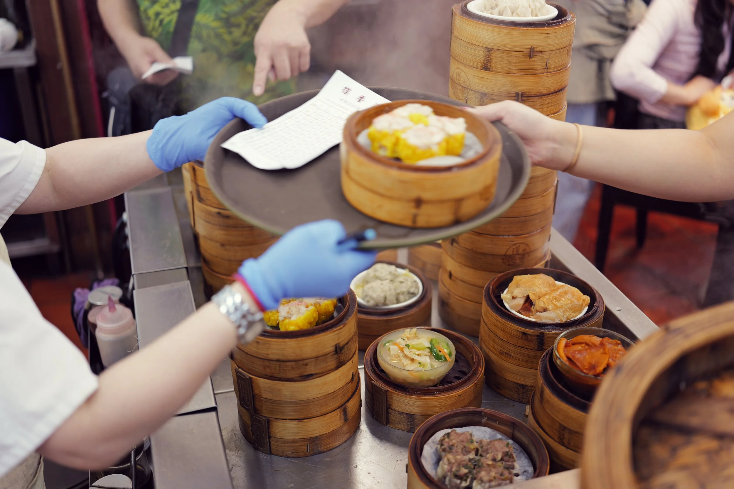 People serving and receiving various dim sum dishes in bamboo steamers at a food stall.