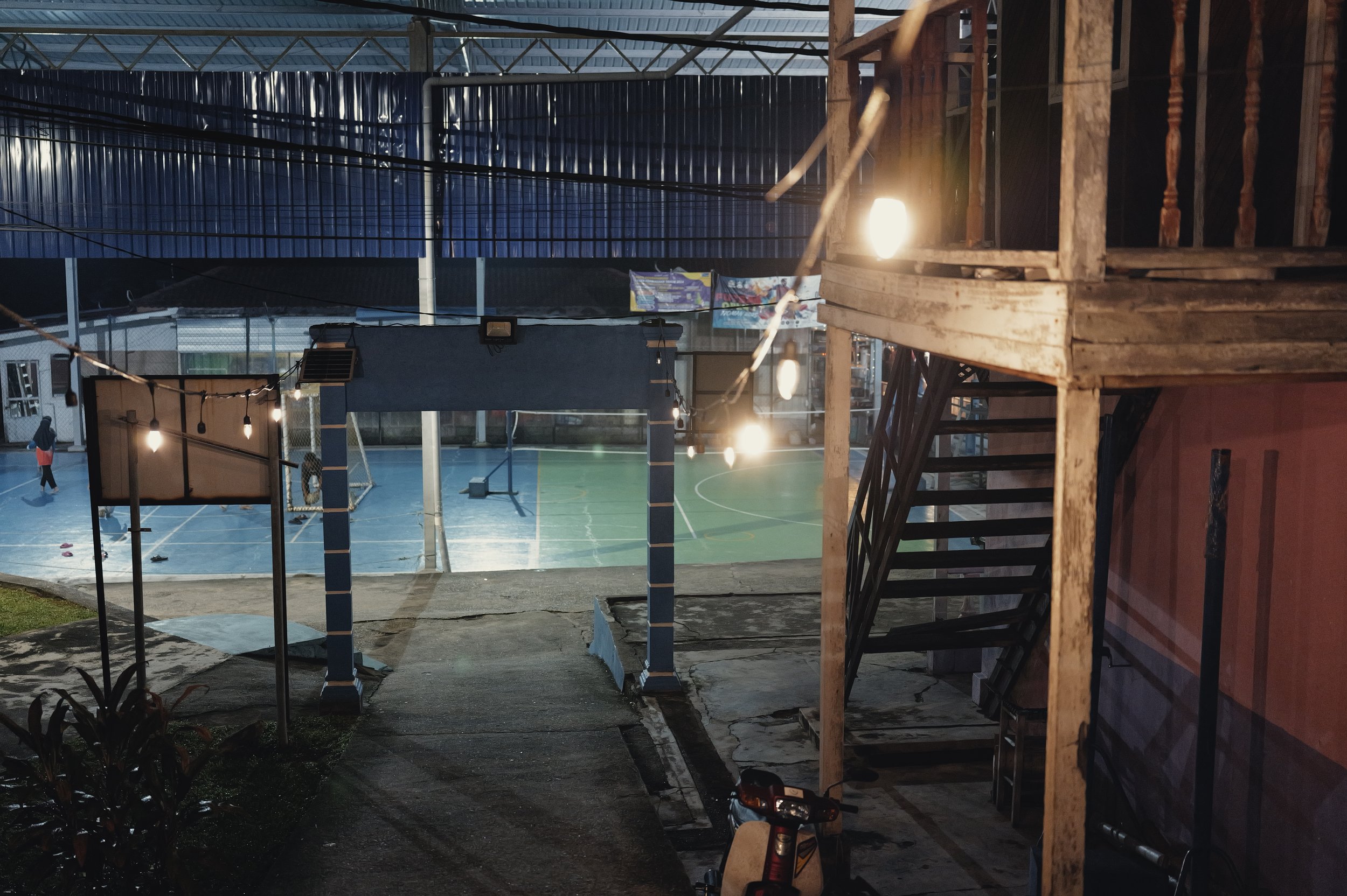 Night view of a sports court, viewed from a balcony with wooden railing, lights illuminating the area, and a person walking on the court.