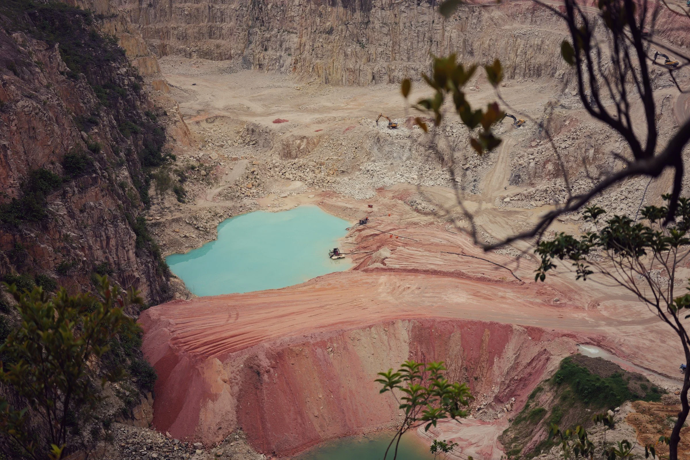 Turquoise quarry water and terraced red earth are framed by branches and leaves.