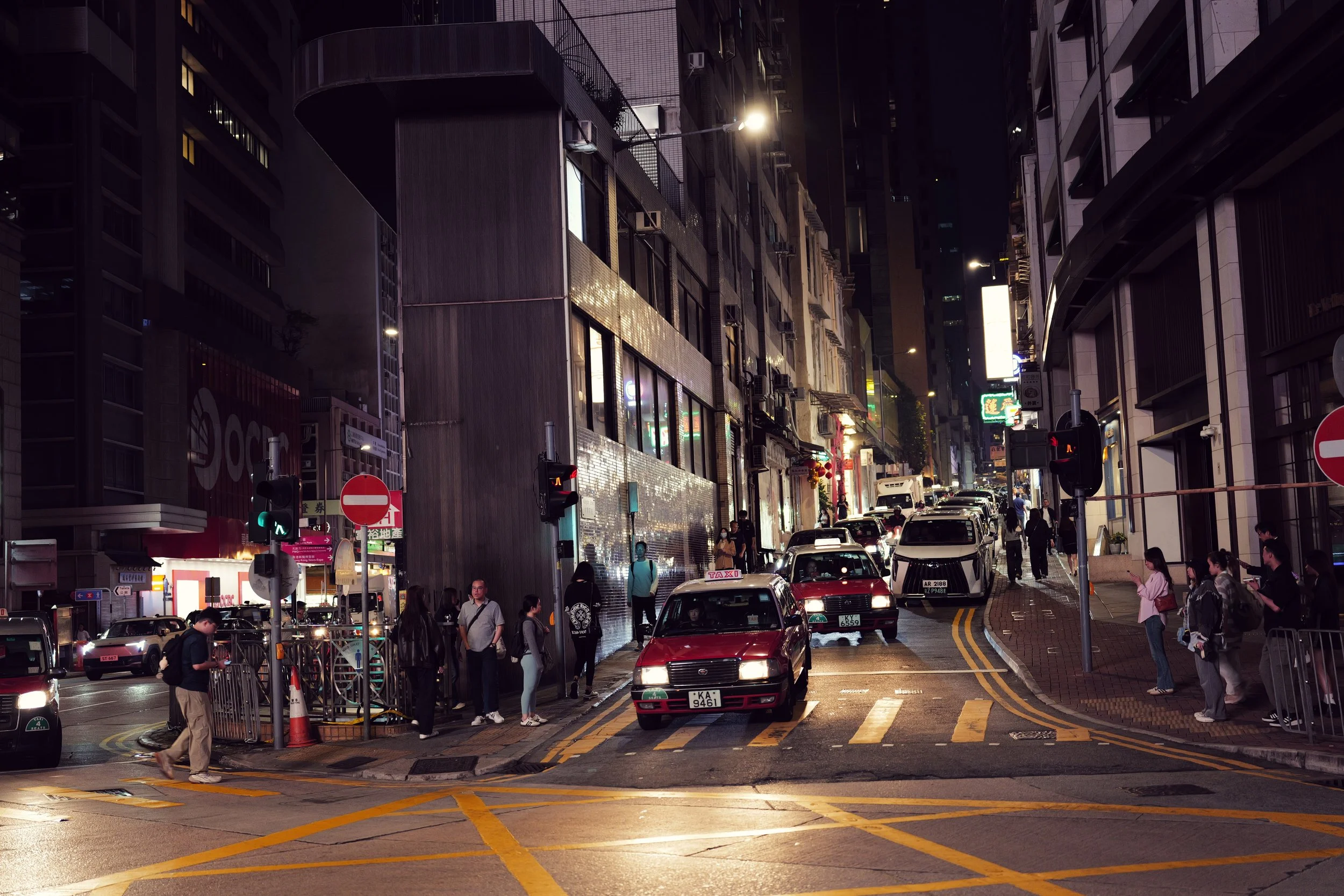 A busy city street at night with people walking and cars stopped at a traffic light. Tall buildings with illuminated signs and windows line the street.