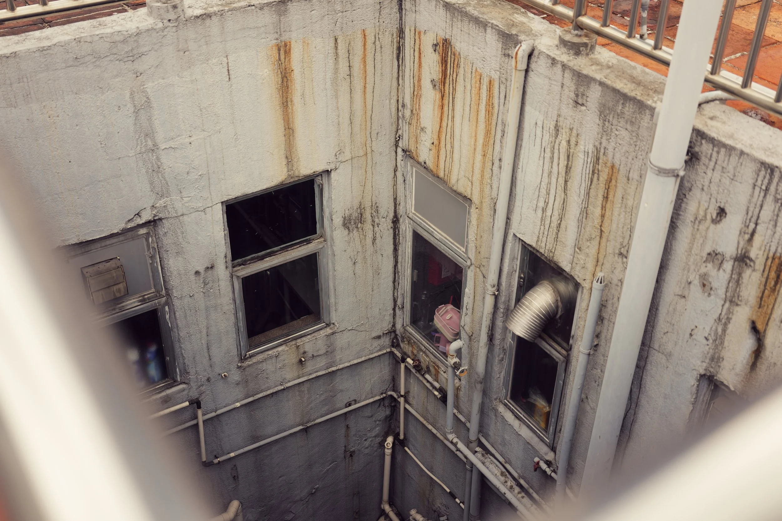 The photo shows a view of an older building's exterior wall with peeling paint, rust stains, and multiple windows. Several pipes and vents run along the wall, and there is a small, pink storage container visible through one window.