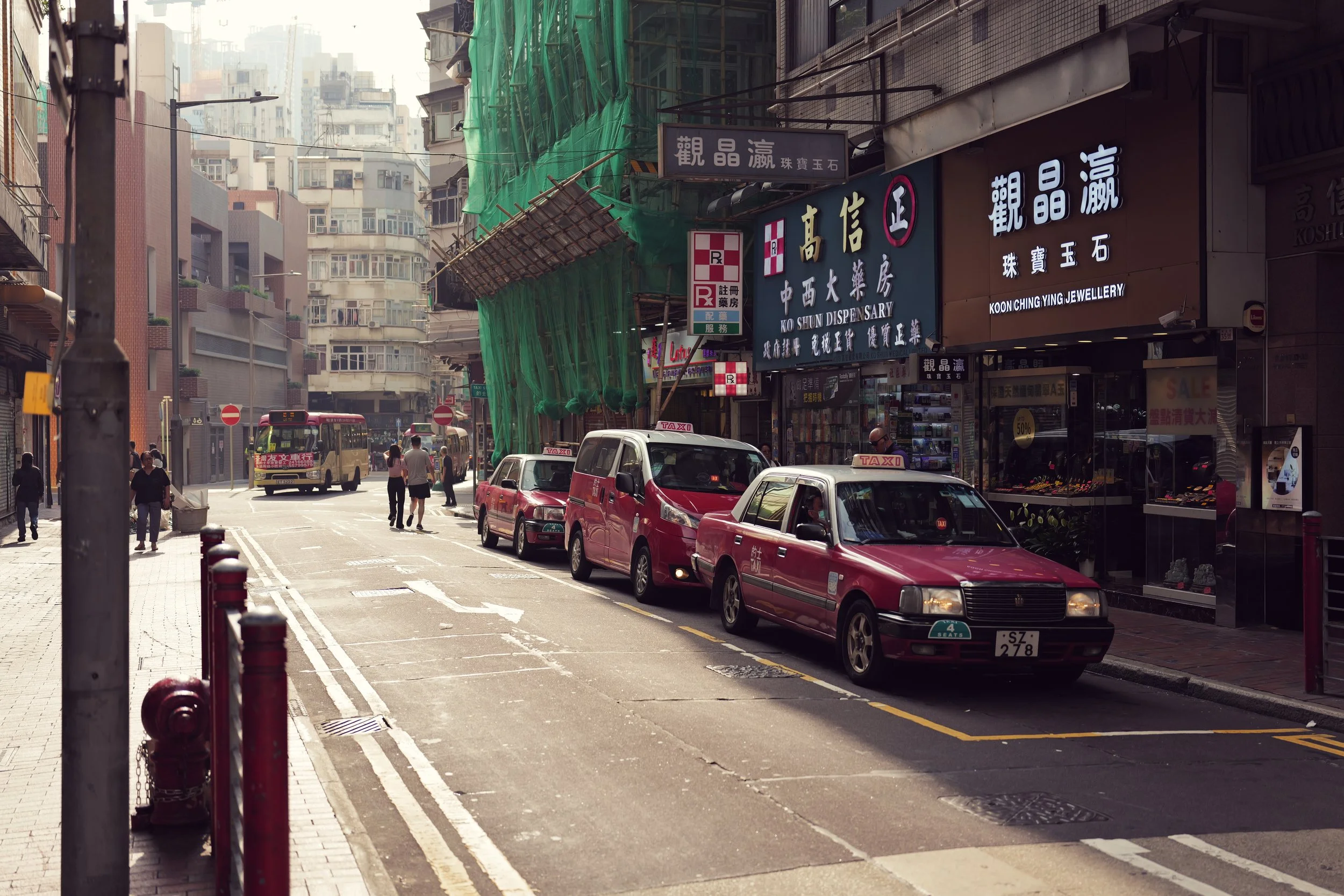 City street scene with pedestrians, parked taxis, storefront signs, and construction covered with green netting.