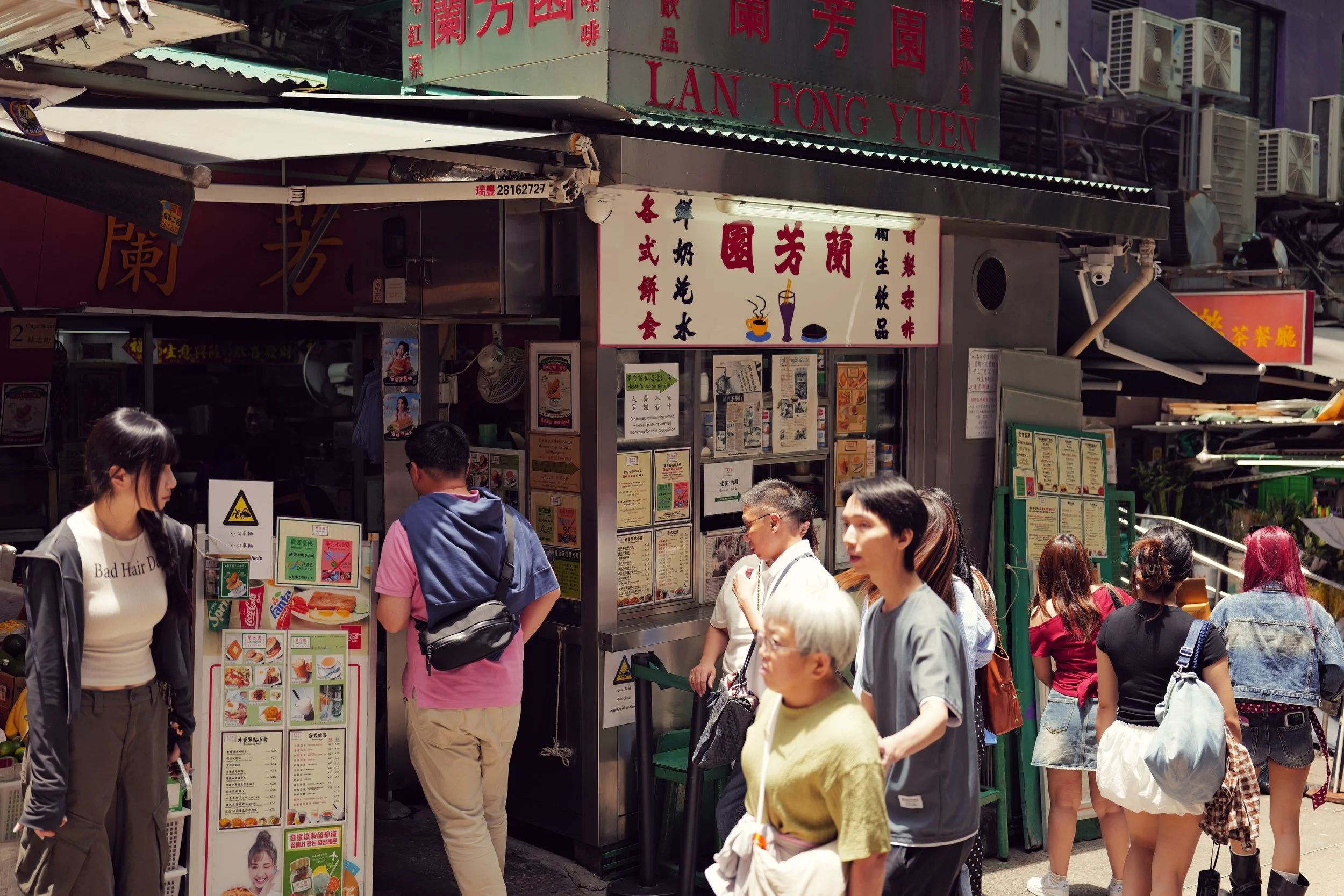 Street scene with multiple people standing and walking in front of food stalls with colorful signs and menus, some signs in Chinese characters.