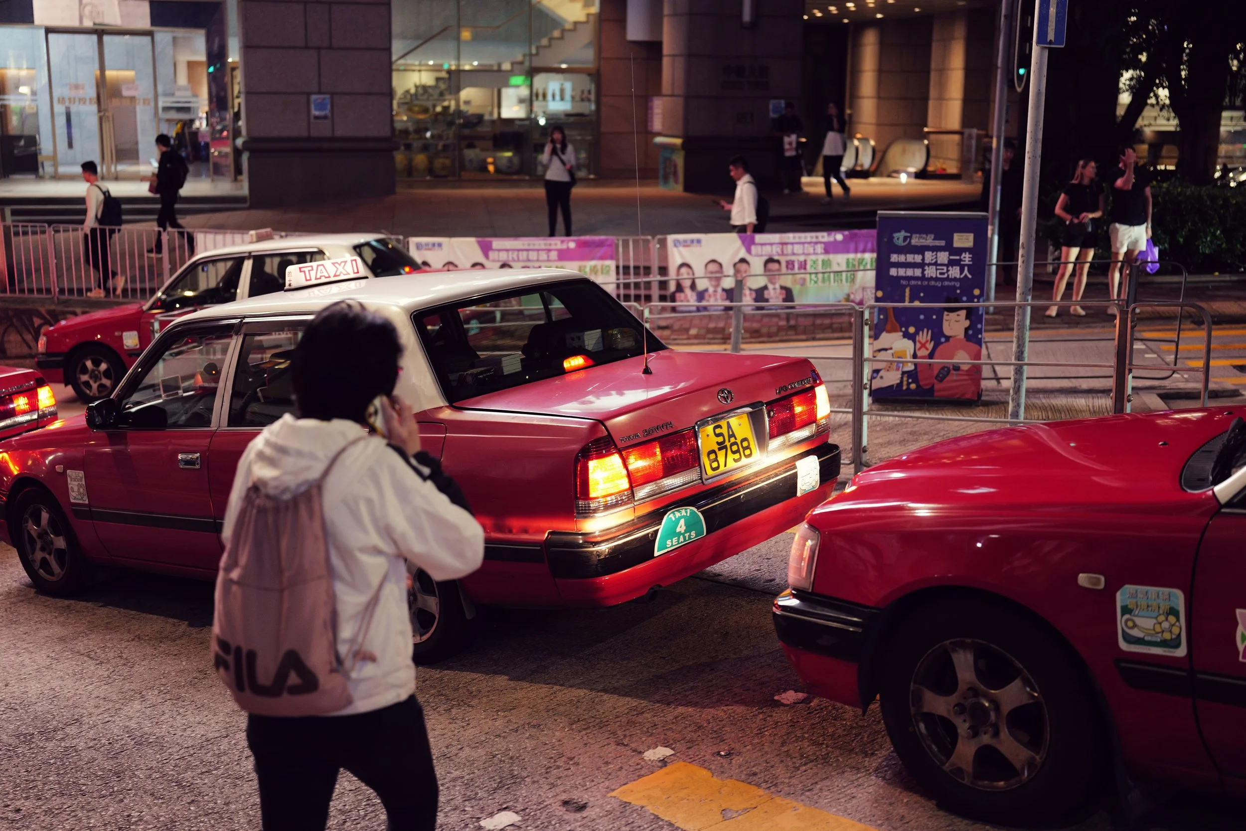 A busy city street scene at dusk with red taxis parked along the curb, pedestrians walking, and a modern building in the background. A woman in the foreground wears a light-colored jacket and mask, talking on a phone, while the taxis display roof sig