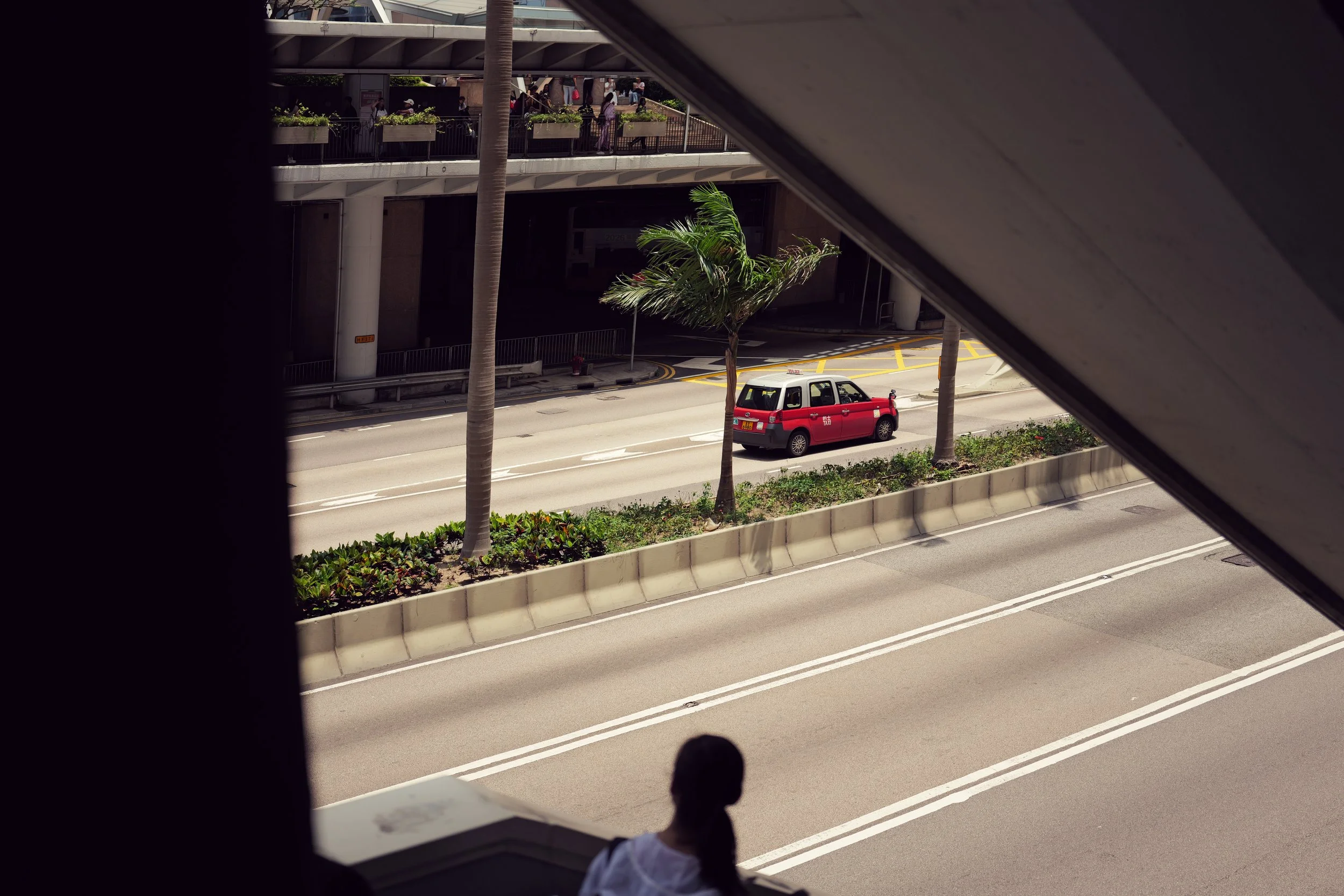 View of city street through an elevated window, with a red taxi driving by and pedestrians on the upper level. Palm trees line the street, and there are concrete barriers separating lanes.