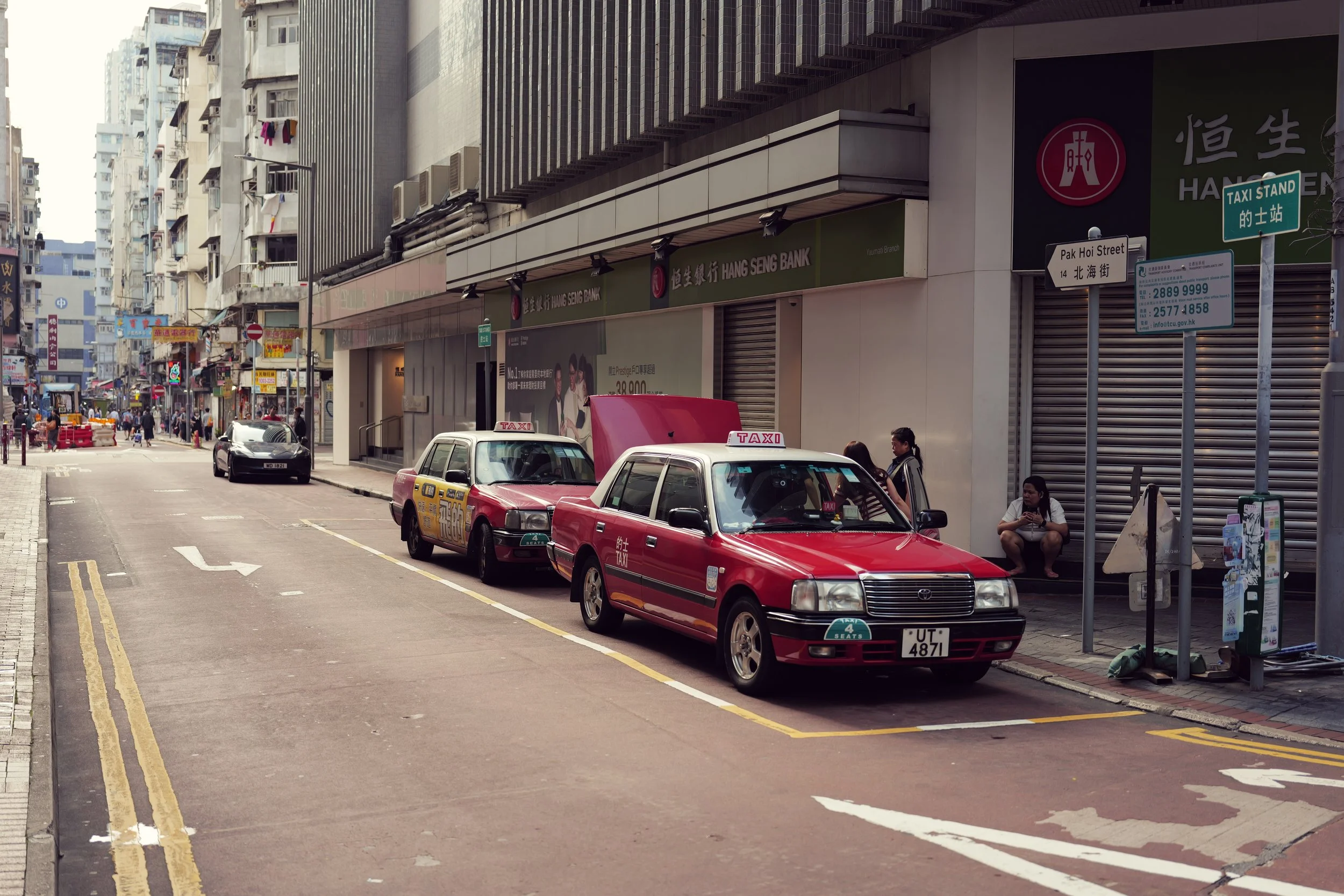 City street scene with parked cars, taxis, people walking and sitting, and buildings with signs including Hang Seng Bank and street signs for Pak Hoi Street and a taxi stand.