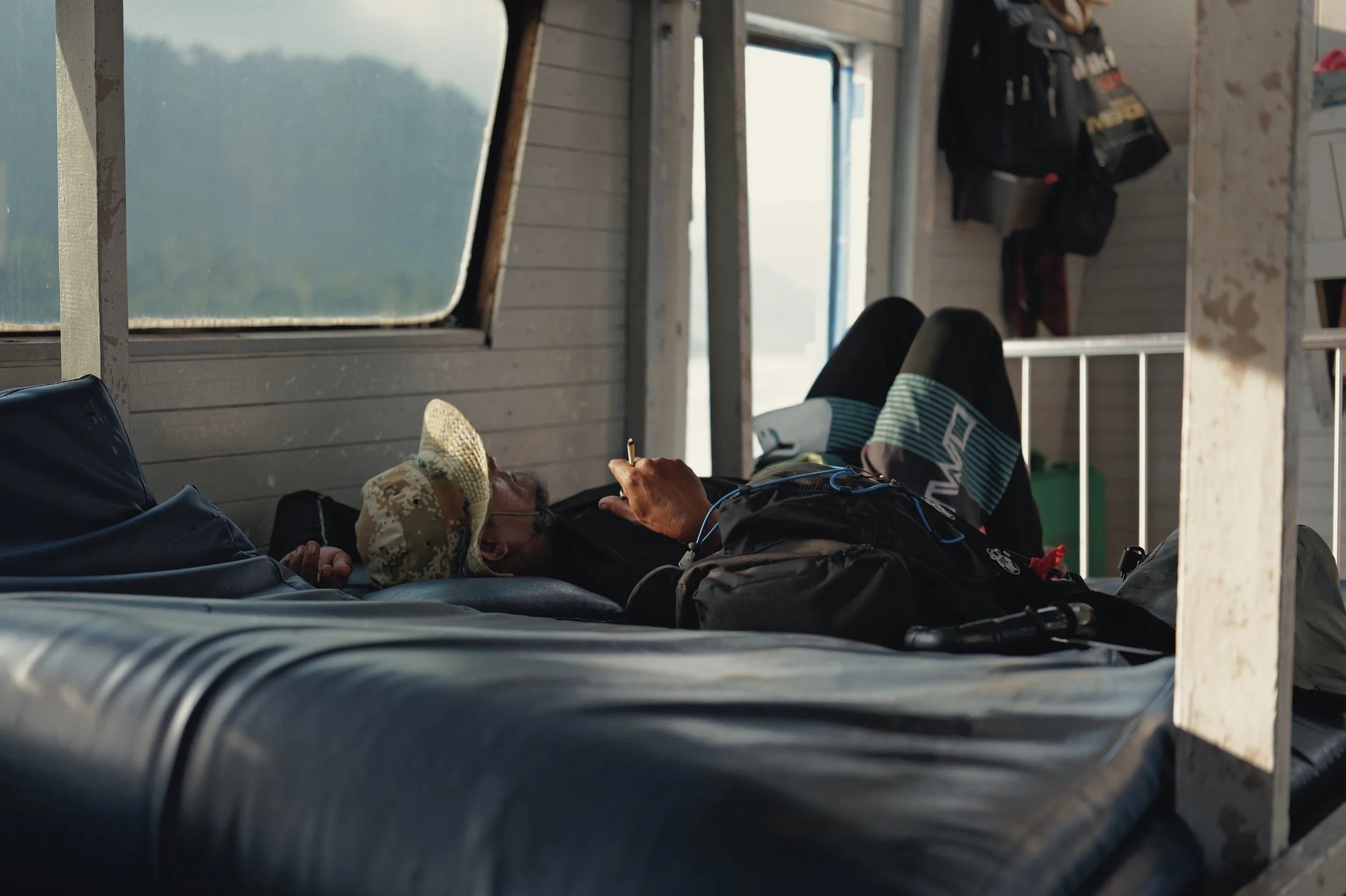 A person lying on a cot inside a rustic wooden structure, wearing a baseball cap and sunglasses, holding a cigarette, with bags and clothing hanging on the wall, and open windows exposing an outdoor landscape.