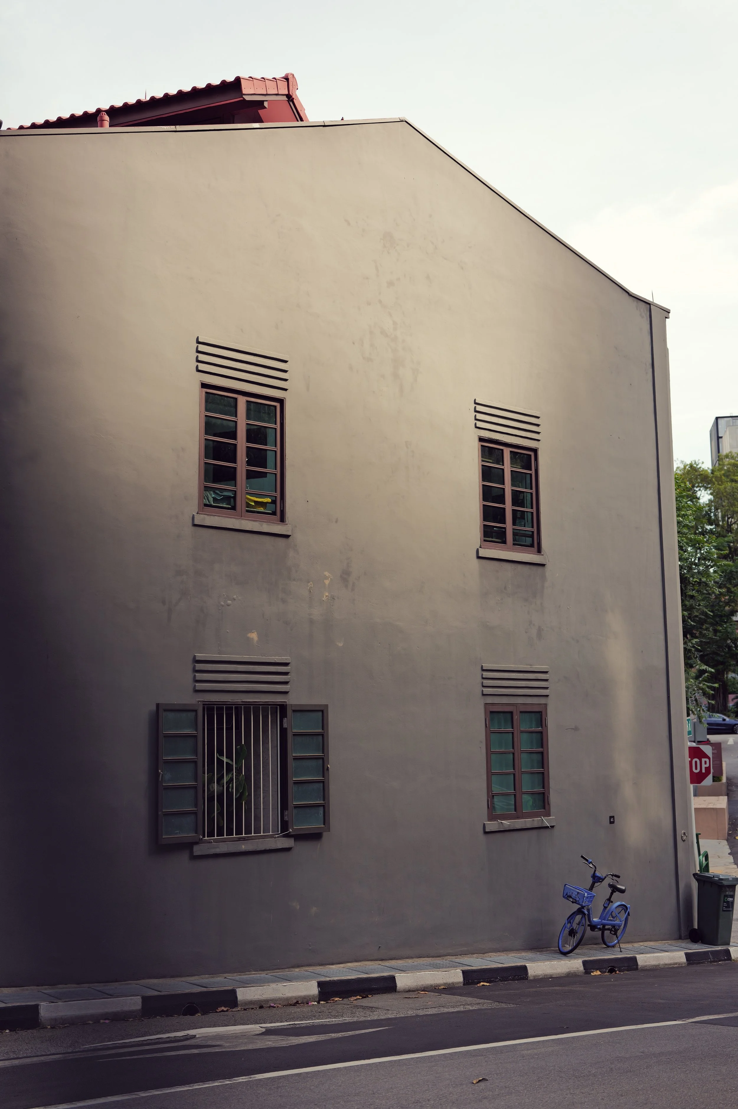 A gray building with several windows, one of which has a plant inside behind bars. There is a blue bicycle parked on the sidewalk and a trash bin nearby. A red stop sign is visible in the background.