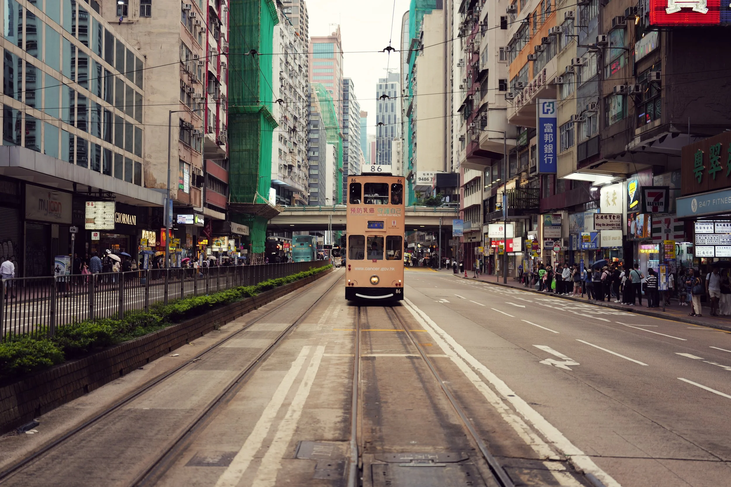 A busy city street with a double-decker tram traveling along tracks, surrounded by tall commercial buildings with shops and signs in various languages.