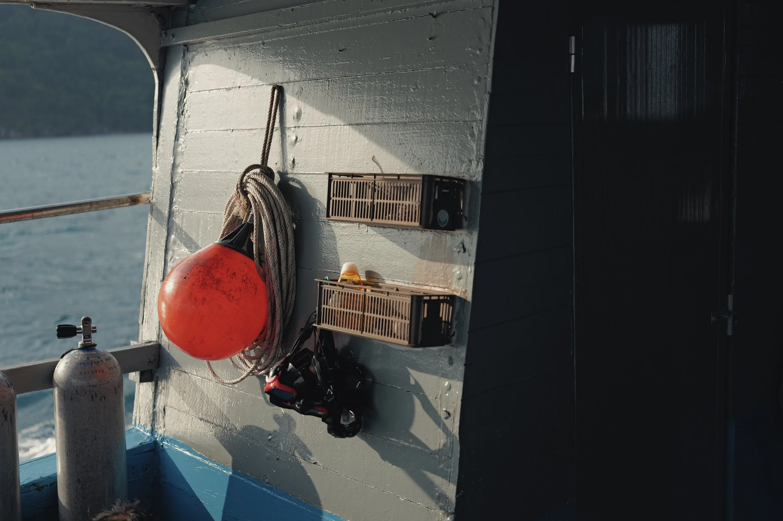 Close-up of a boat's interior showing a fire extinguisher, coiled rope, a small speaker, and two propane tanks, with water and mountains visible through an opening.