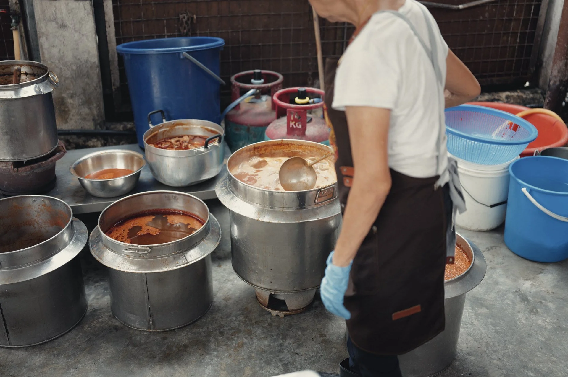 A hawker preparing Penang prawn noodles beside large broth pots and cooking utensils.