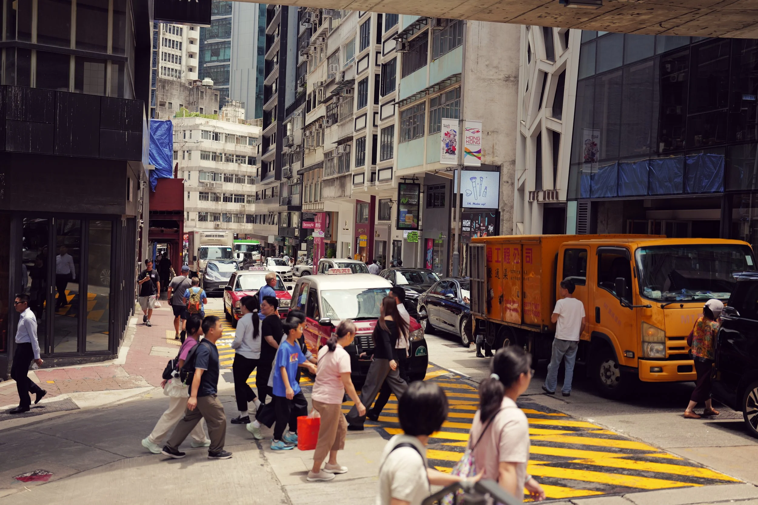 A busy city street corner with pedestrians crossing, cars and trucks on the road, and tall buildings in the background.