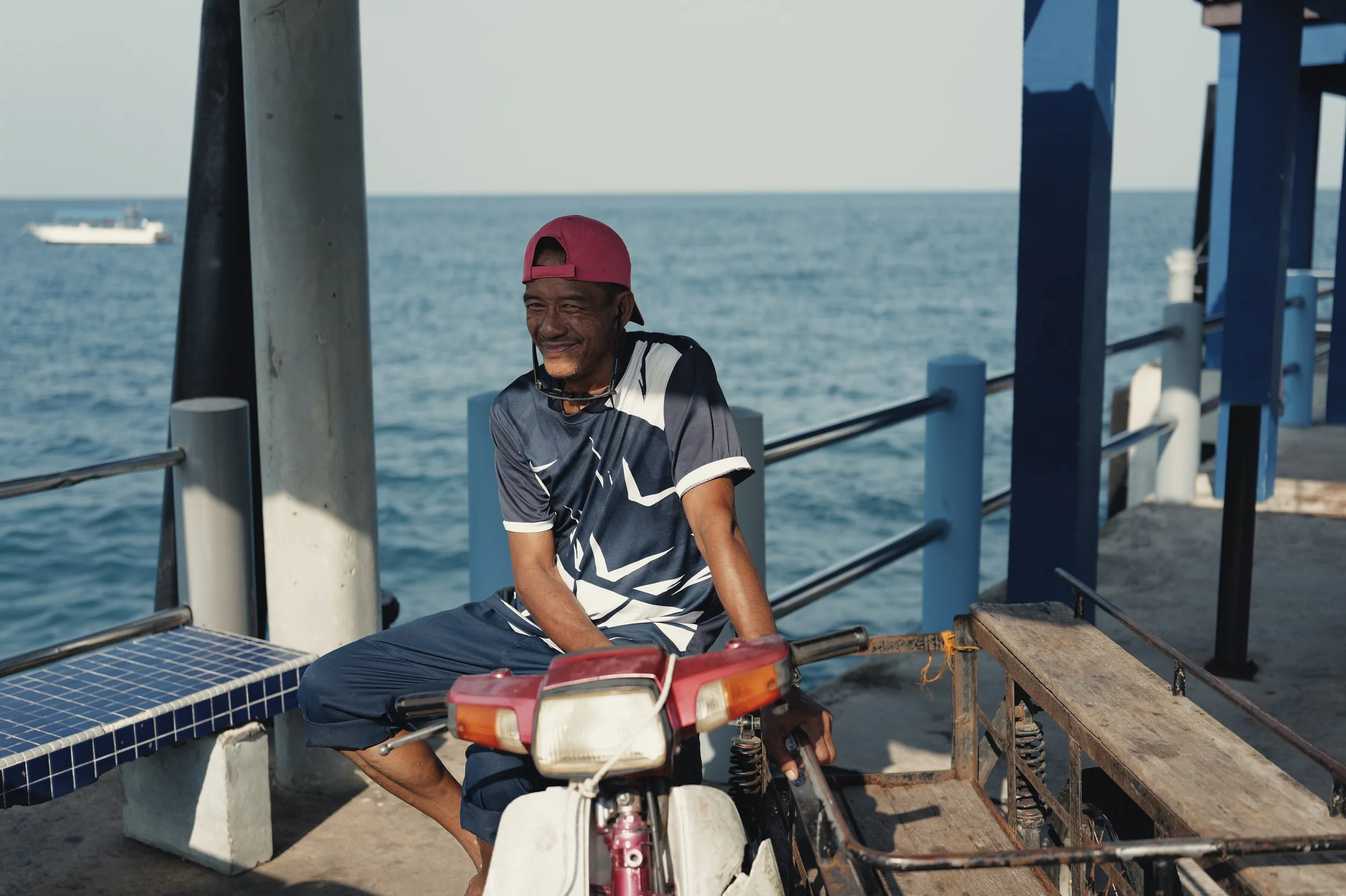 An older man wearing a pink baseball cap and a black and white sports shirt sits on a bicycle near the ocean.