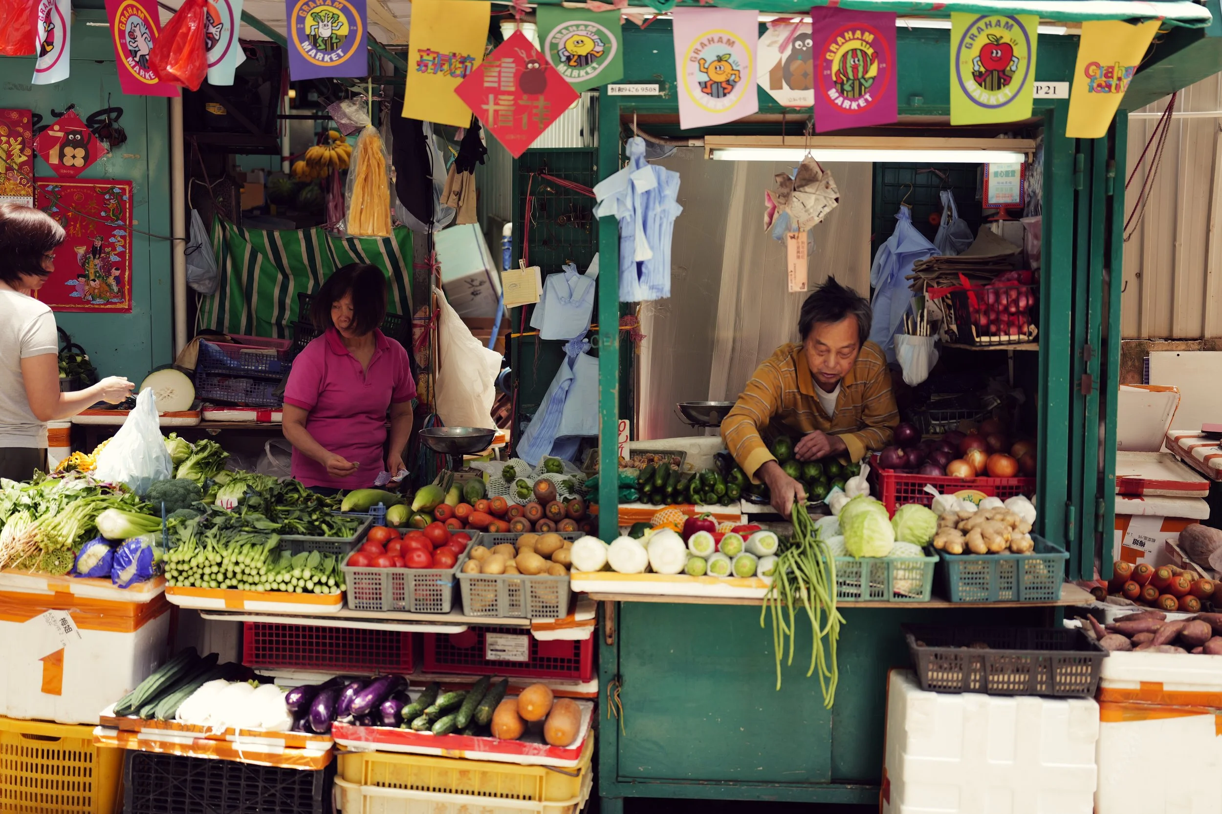 A street market stall displaying fresh vegetables such as cucumbers, tomatoes, eggplants, onions, and leafy greens, with colorful banners and signs in the background.