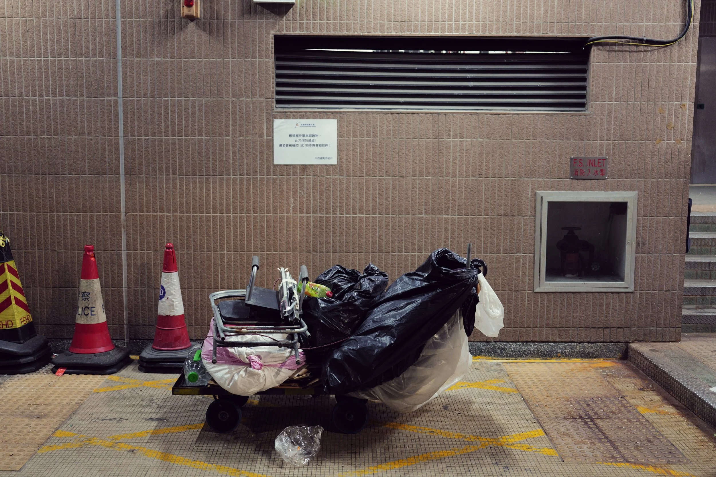A shopping cart filled with black trash bags, placed in front of a brown brick wall with warning and notices, surrounded by traffic cones and a yellow caution line on the ground.