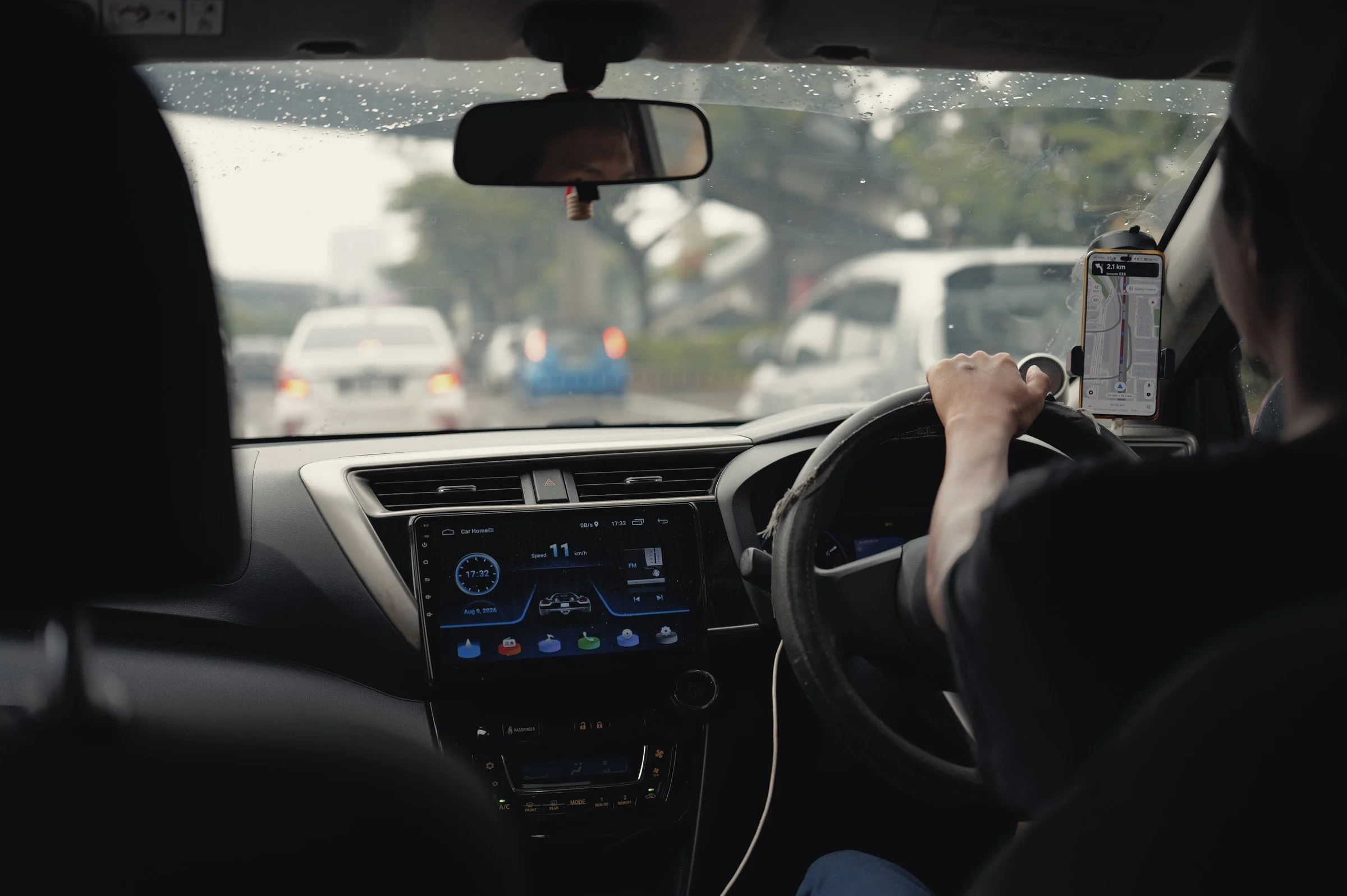 The road ahead is seen from inside a car, past the driver and dashboard.