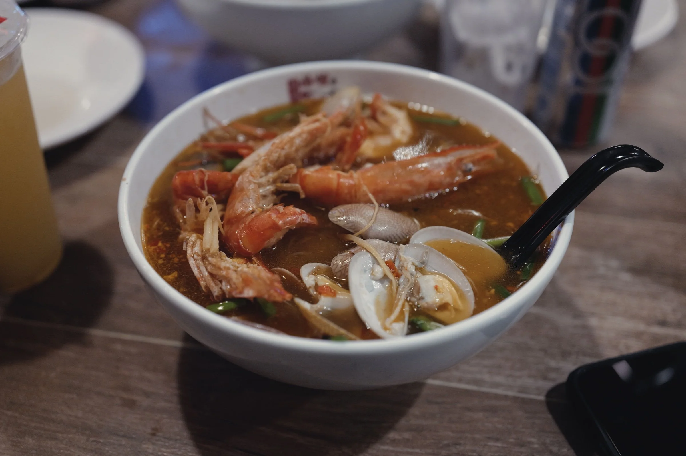 Prawns and clams fill a bowl of reddish seafood noodle soup.