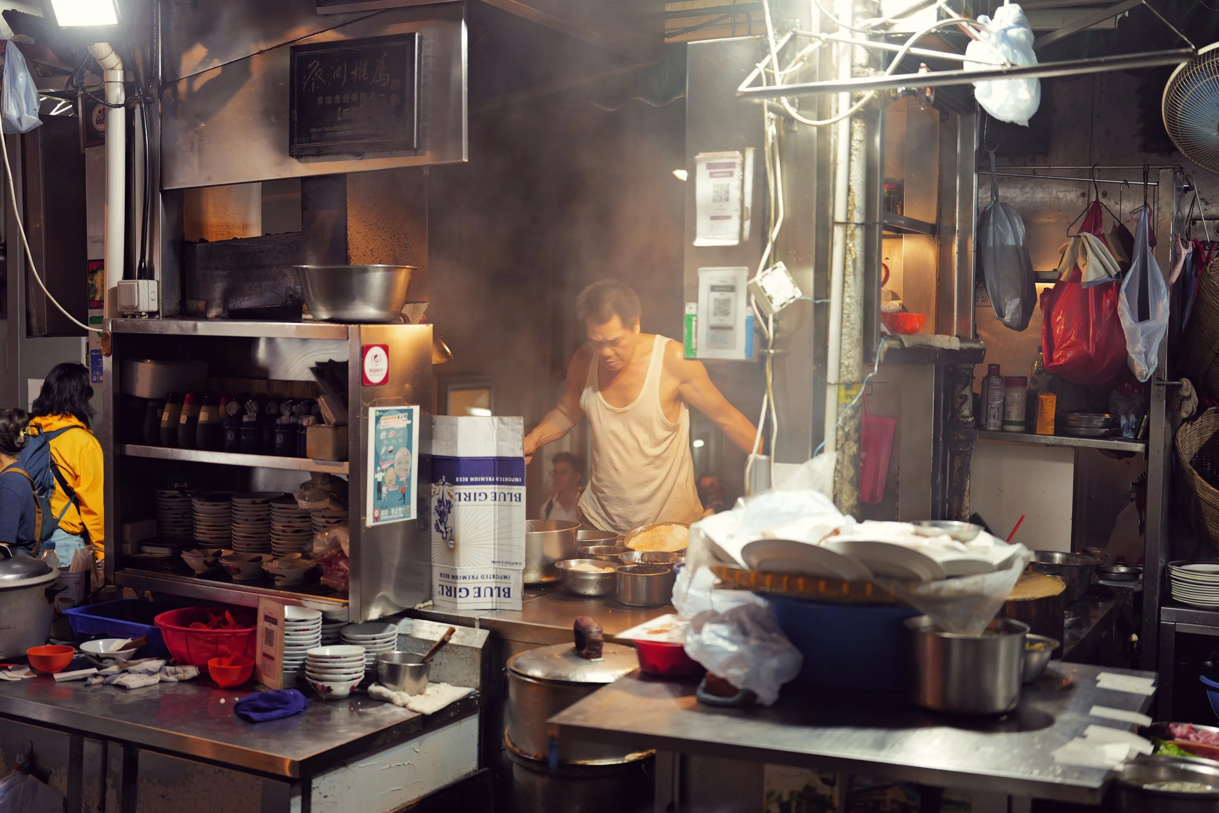 A lively street food stall with a cook preparing dishes behind the counter. There are various bowls, utensils, and ingredients scattered on the counter, with people waiting in line. Steam rises from the cooking area, creating an active and bustling a