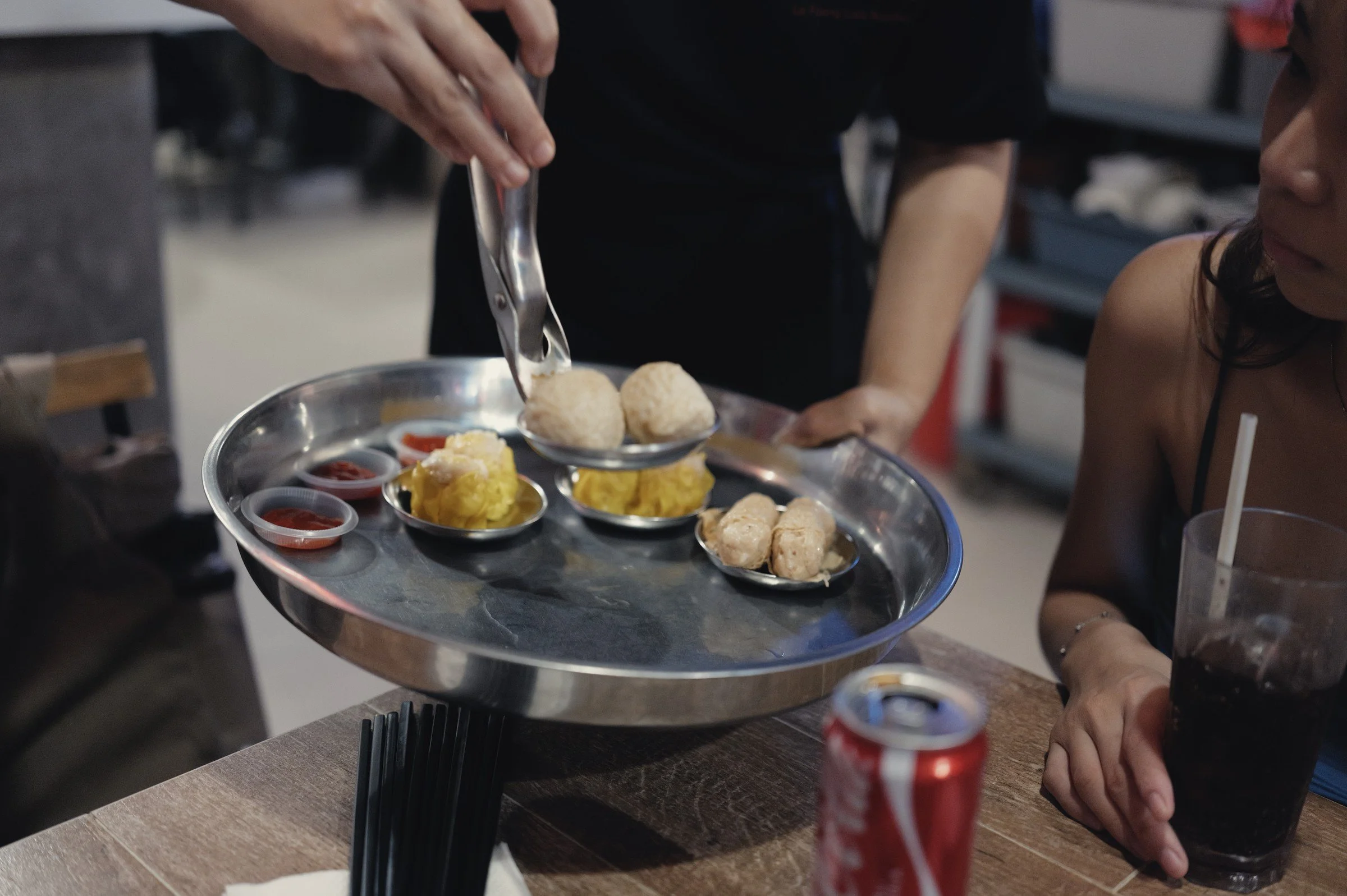 A hand uses tongs to serve dim sum from a round metal tray at a dining table.