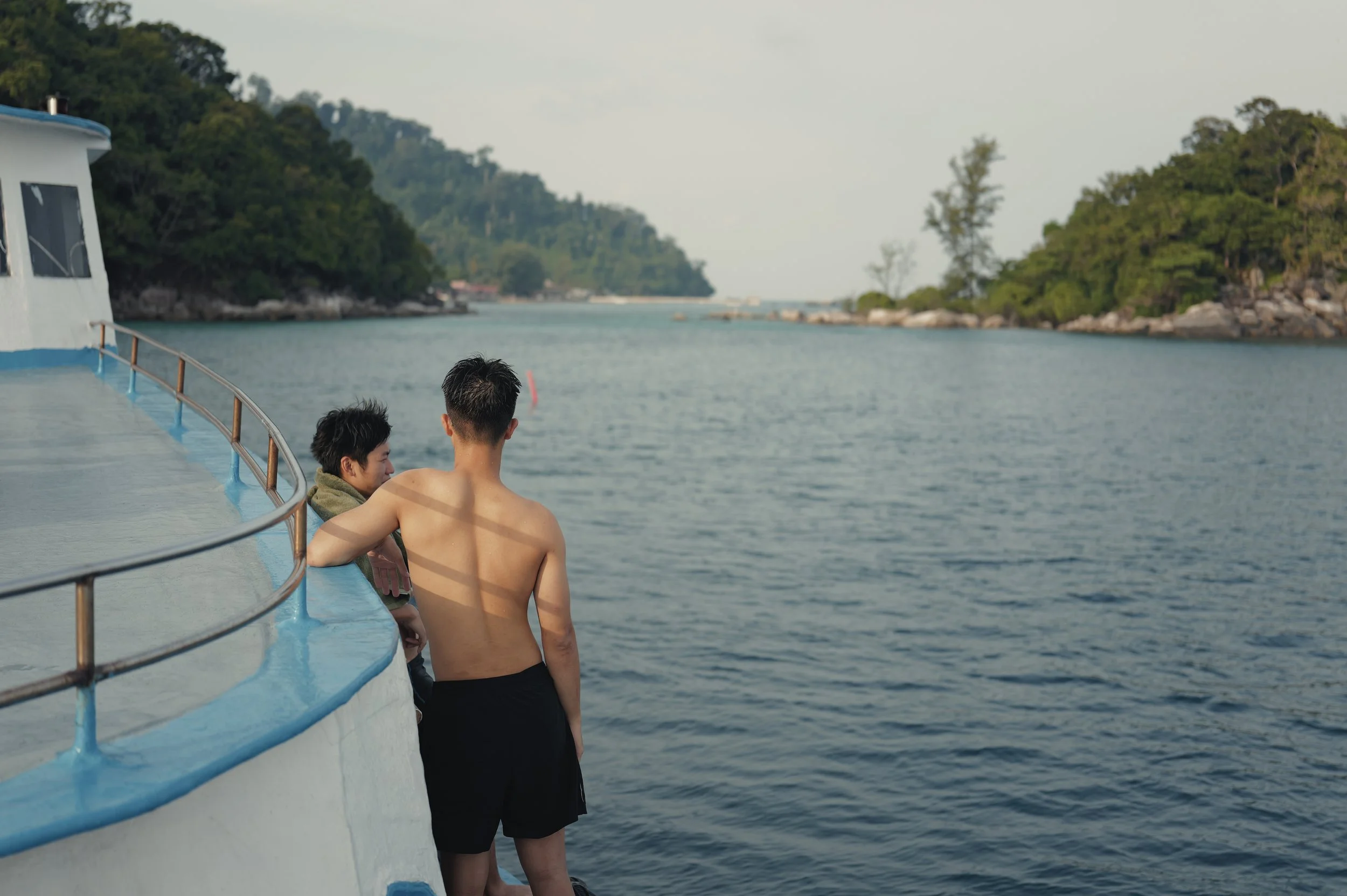 Two young men standing on the deck of a boat, looking at the water and surrounding green island landscape.