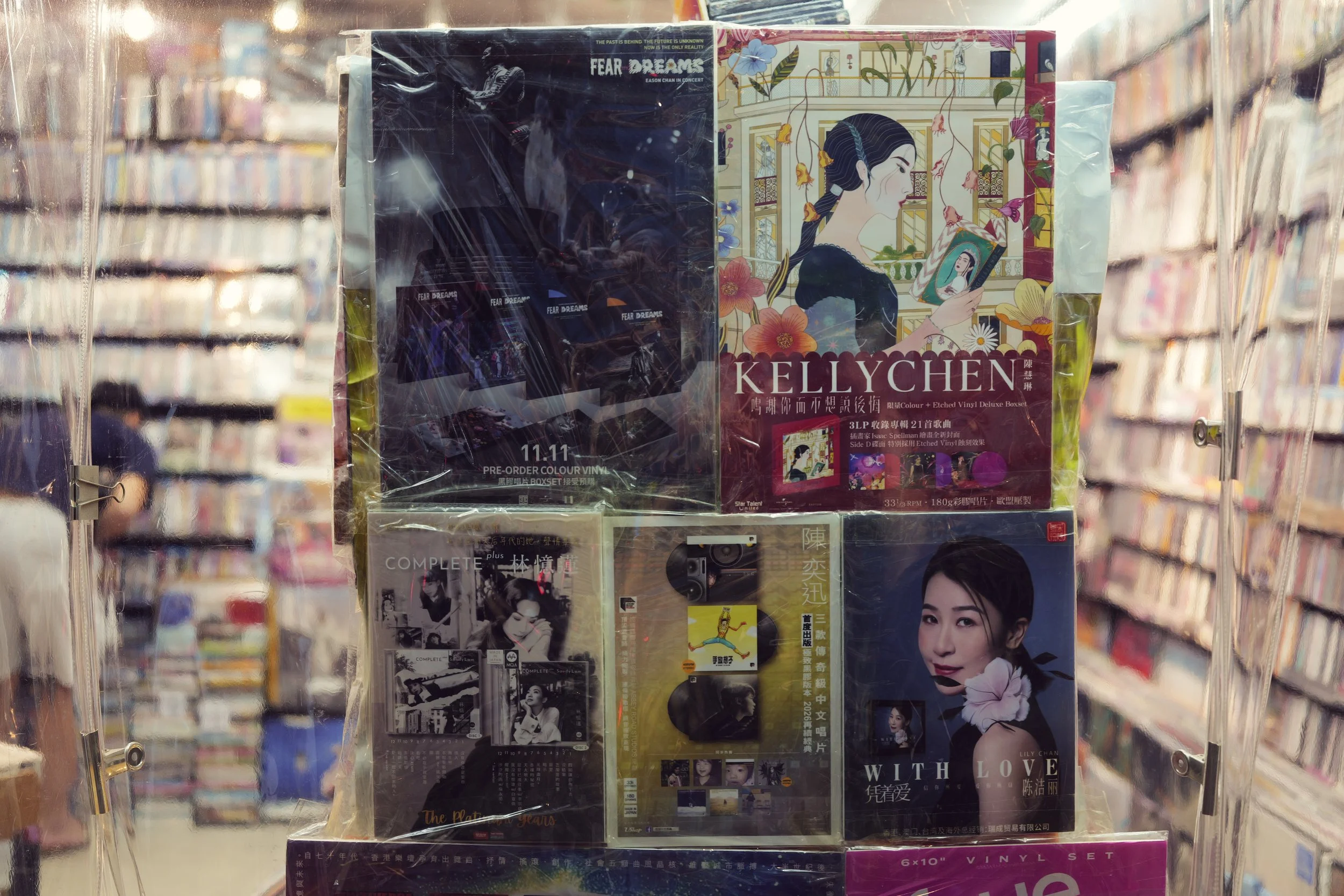 Close-up of a display of vinyl records, including an album titled 'Kelly Chen' featuring an illustration of a woman holding a phone, with additional albums with Asian scripts and images of women visible behind. The display is behind a clear plastic c