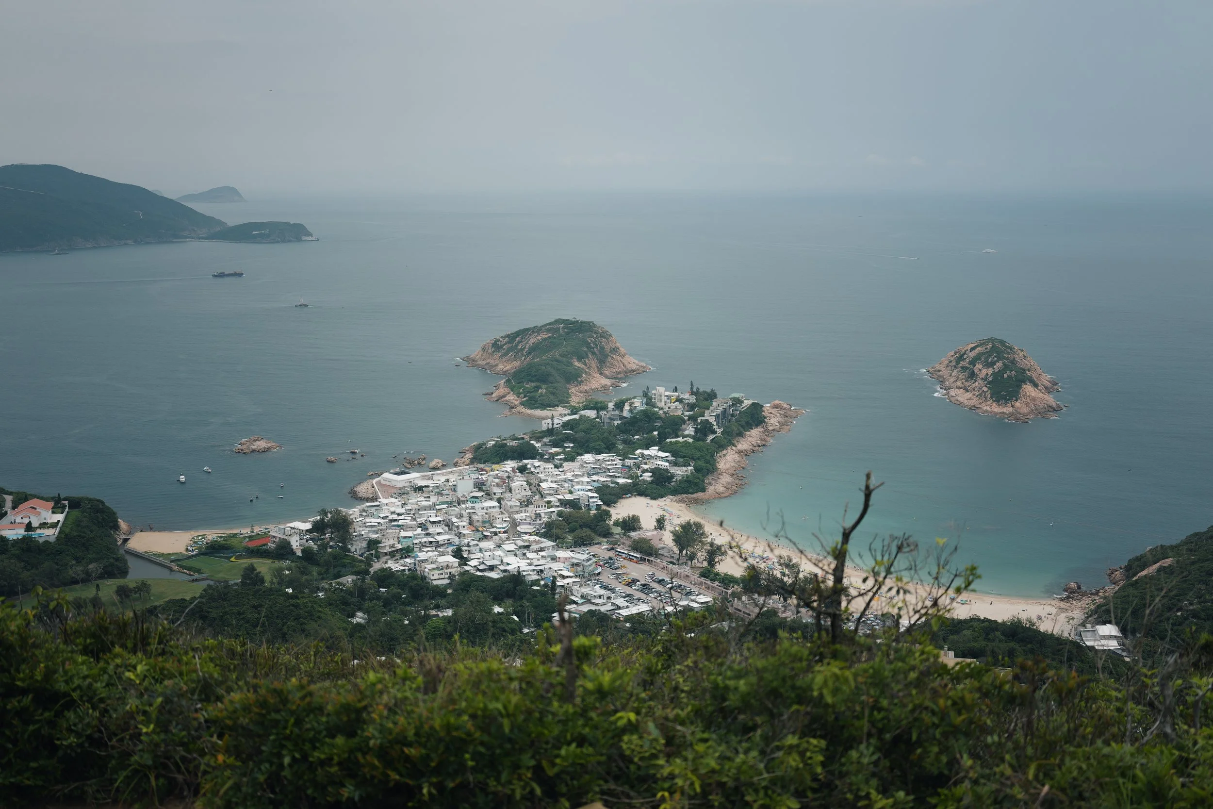 A coastal town with white buildings along the shoreline, small islands in the water, and boats sailing across a calm sea, viewed from a hillside with green foliage.