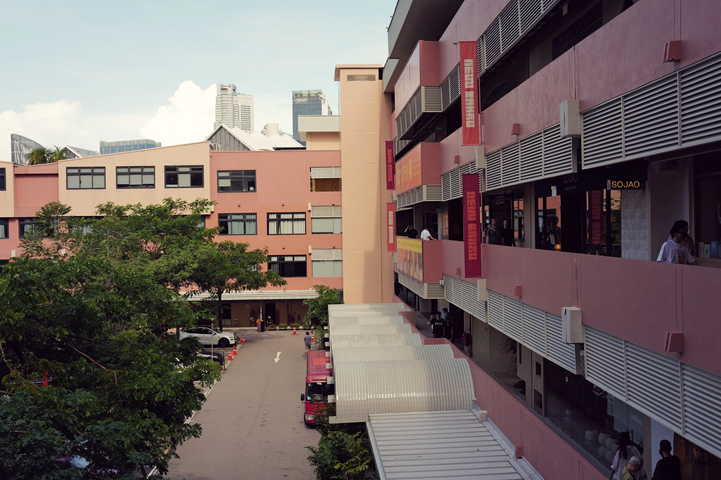 A pink commercial building with retail shops and people walking on the sidewalk, bordered by trees and parked cars, with a city skyline in the background.