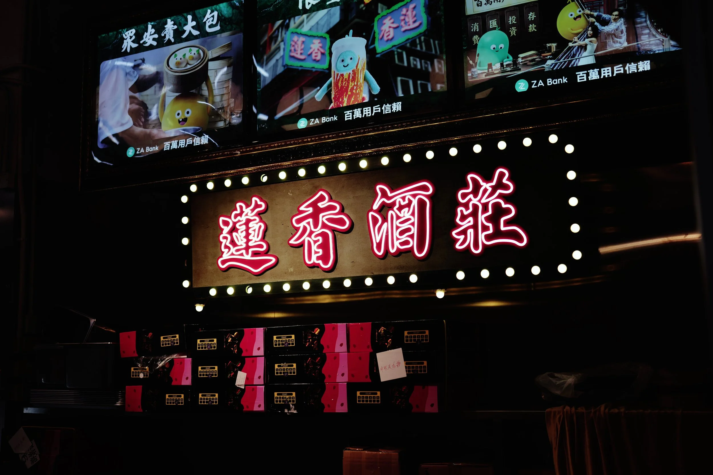 Bright neon signs with Chinese characters and animated characters including a smiling yellow ball, a cartoon cupcake, and a green creature, displayed above a store in an urban setting at night.