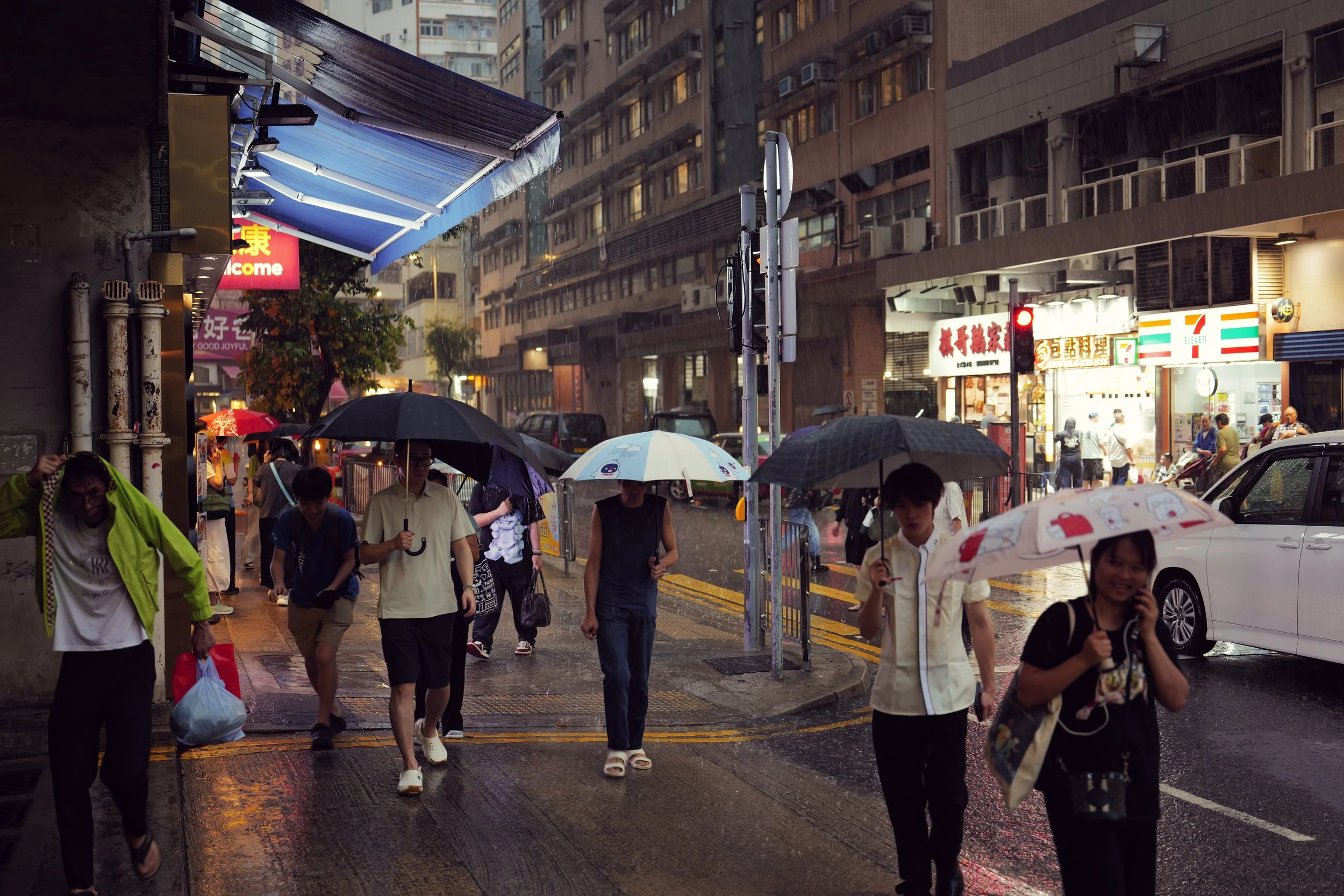 People walking on a rainy city street with umbrellas, in front of a storefront with Asian signage and a 7-Eleven, with wet pavement reflecting city lights.